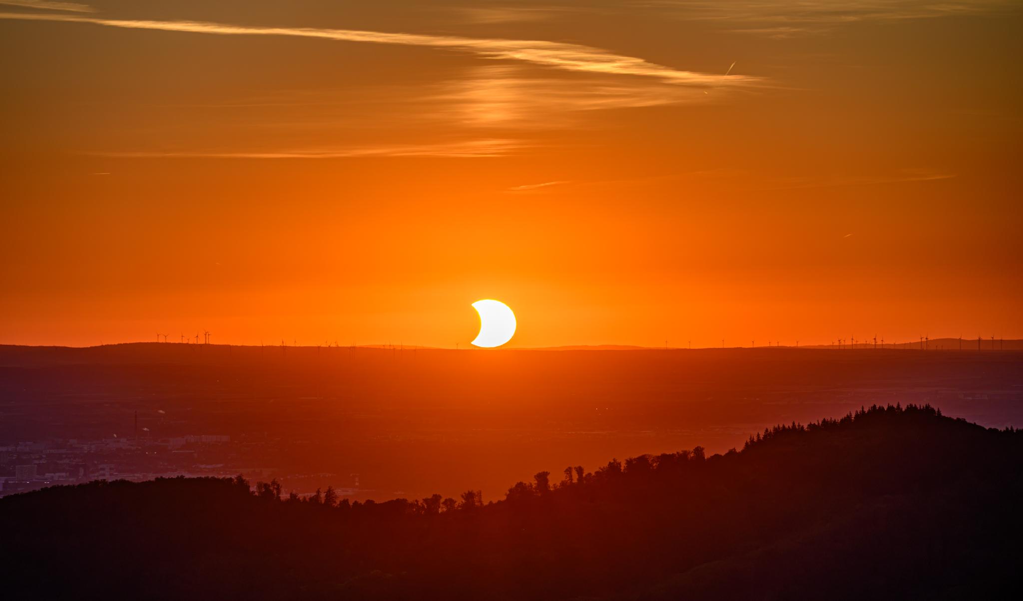 The Sun just about to touch the western horizon at sunset, partially eclipsed by the Moon on the left-hand side, taking a bite out of it. The sky is yellow and orange, and there are many windmills in the distance along the hills of the Pfalz on the other side of the Rhine plain. In the middle distance, the plain can be seen with part of Mannheim, while in the foreground, the foothills of the Odenwald are in darker shades. There are some wisps of clouds in the sky above the Sun.