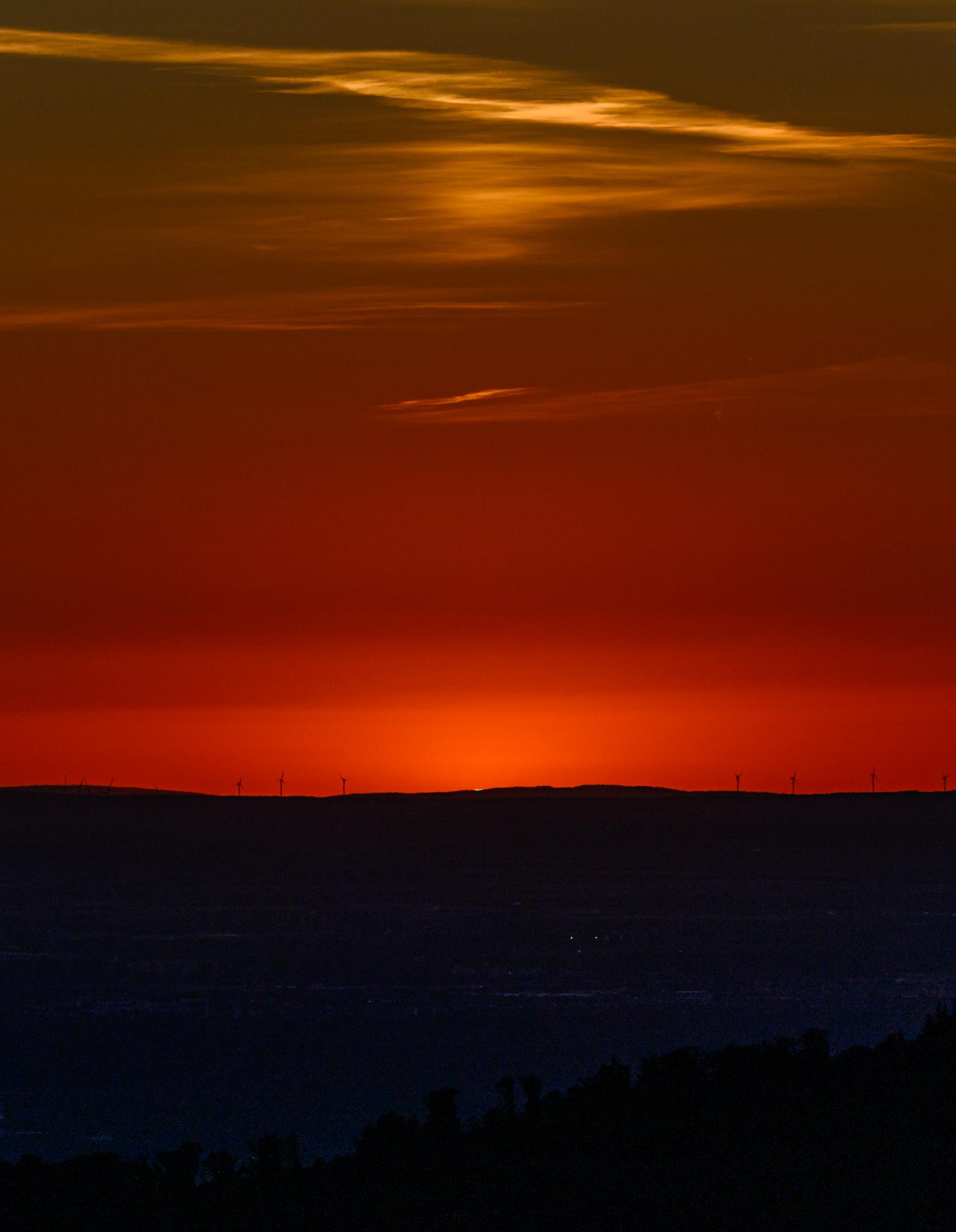 A horizon of hills right at the end of sunset. The hills are dark against a red sky, with some wisps of yellow clouds above. There are windmills along the top of the hills. The very last light from the setting sun is visible, showing a distinct green colour.