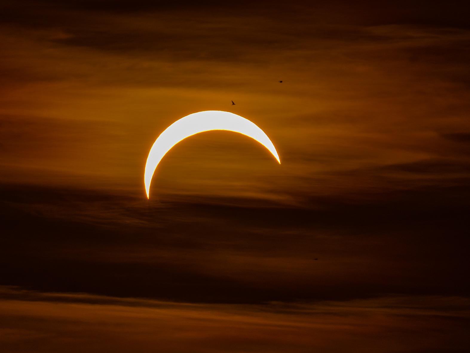 Ein oranger leicht bewölkter Himmel. Im Zentrum die Sichel der Sonnenscheibe, von der rechts unten ein großer Teil fehlt