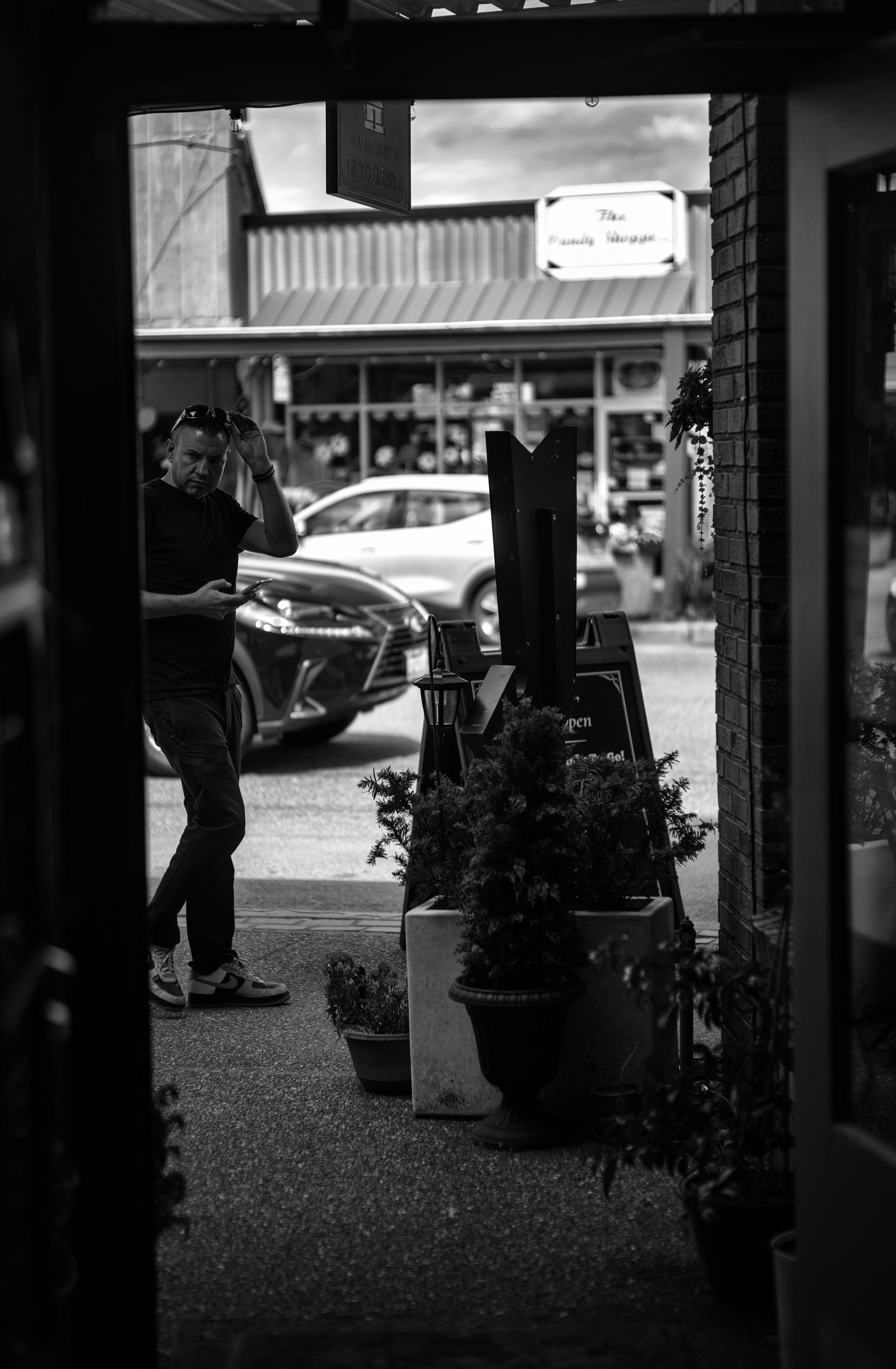 Black and white photo in portrait format of a man caught in mid-stride walking outside of a small business.

The photo was taken inside the business looking out into the street through the front entrance to create a slightly left-tilted "frame". A male passerby, dressed in dark pants and short sleeve T-shirt, is entering the frame from the left and looking at something on the outside wall facing the street. His left hand is in the middle of pulling down a pair of sunglasses propped on his head. His right arm is bent at a 90 degree angle at his side, and he's holding a mobile phone with thumb above it's face as if in the middle of texting someone.

Outside the business, lined on the sidewalk are a number of small and large planters visible in the middle of the door frame. In the far background two cars passing by in opposite directions are partially visible, as is another small storefront on the opposite side of the street.