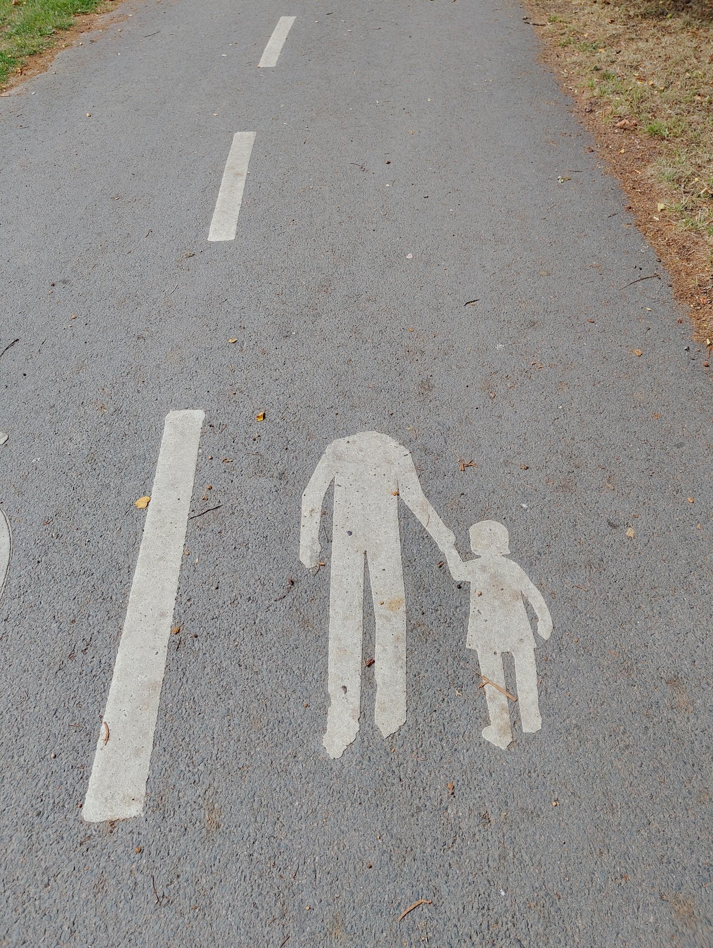 Auf einem asphaltierten Rad-und Fußweg ist neben einem Trennstrich der beiden Seiten ein Fußgängersymbol durch weißen Asphalt aufgebracht: Es zeigt eine erwachsene Person mit einem Kind an der Hand. Bei der erwachsenen Person fehlt allerdings der Kopf.