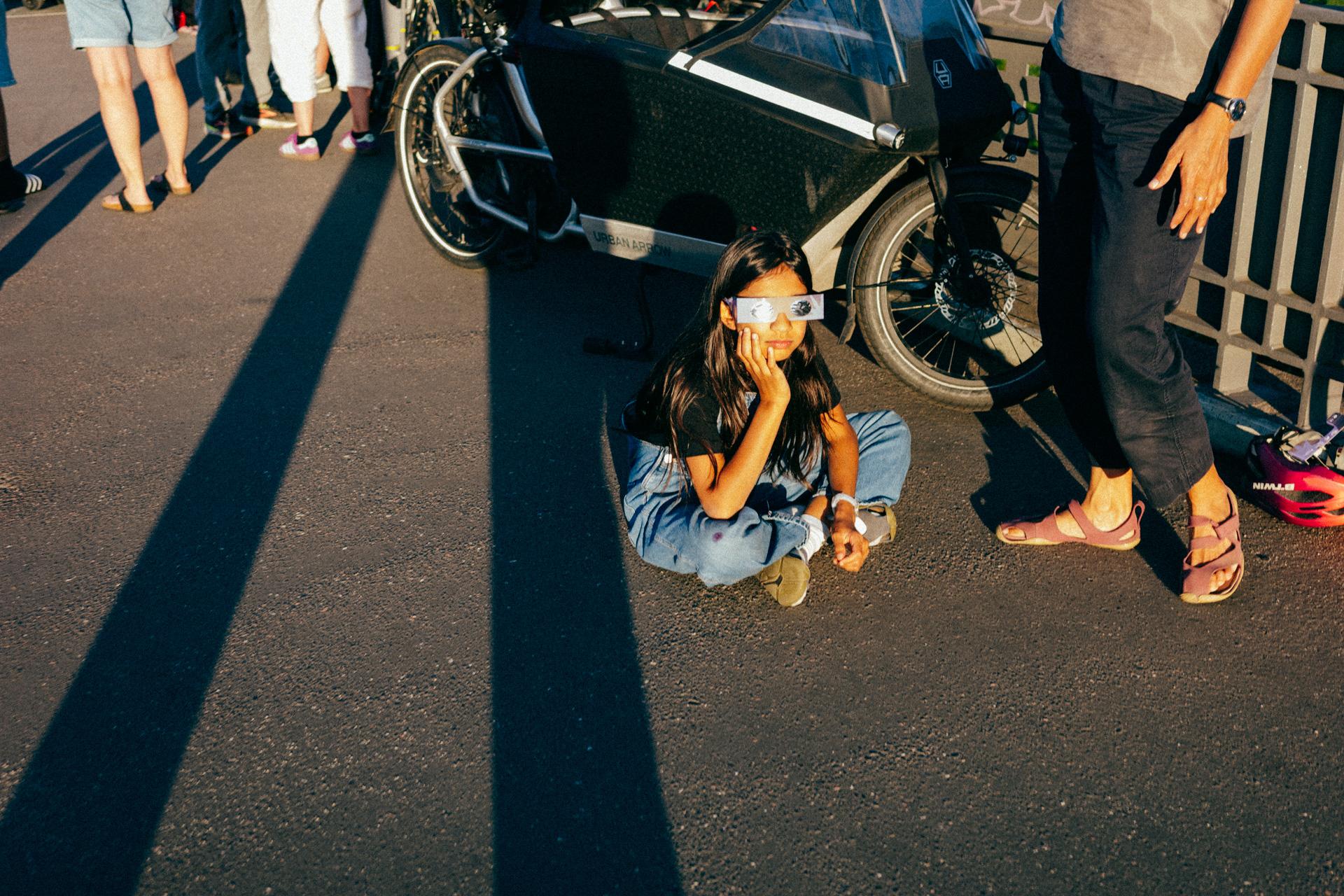 Una niña sentada en el suelo, con cara aburrida y gafas para ver el eclipse.
A girl sitting on the ground, looking bored and wearing eclipse glasses.
