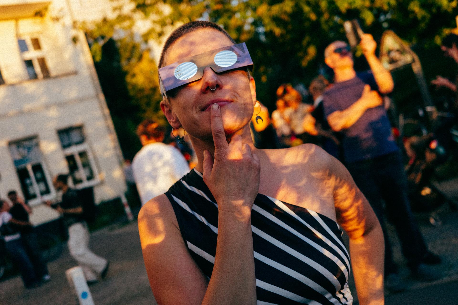 Retrato de una persona con gafas del eclipse. La sombra de los árboles proyecta lunas en su cuerpo y rostro.
Portrait of a person wearing eclipse glasses. The shadow of the trees casts crescent shapes onto their body and face.