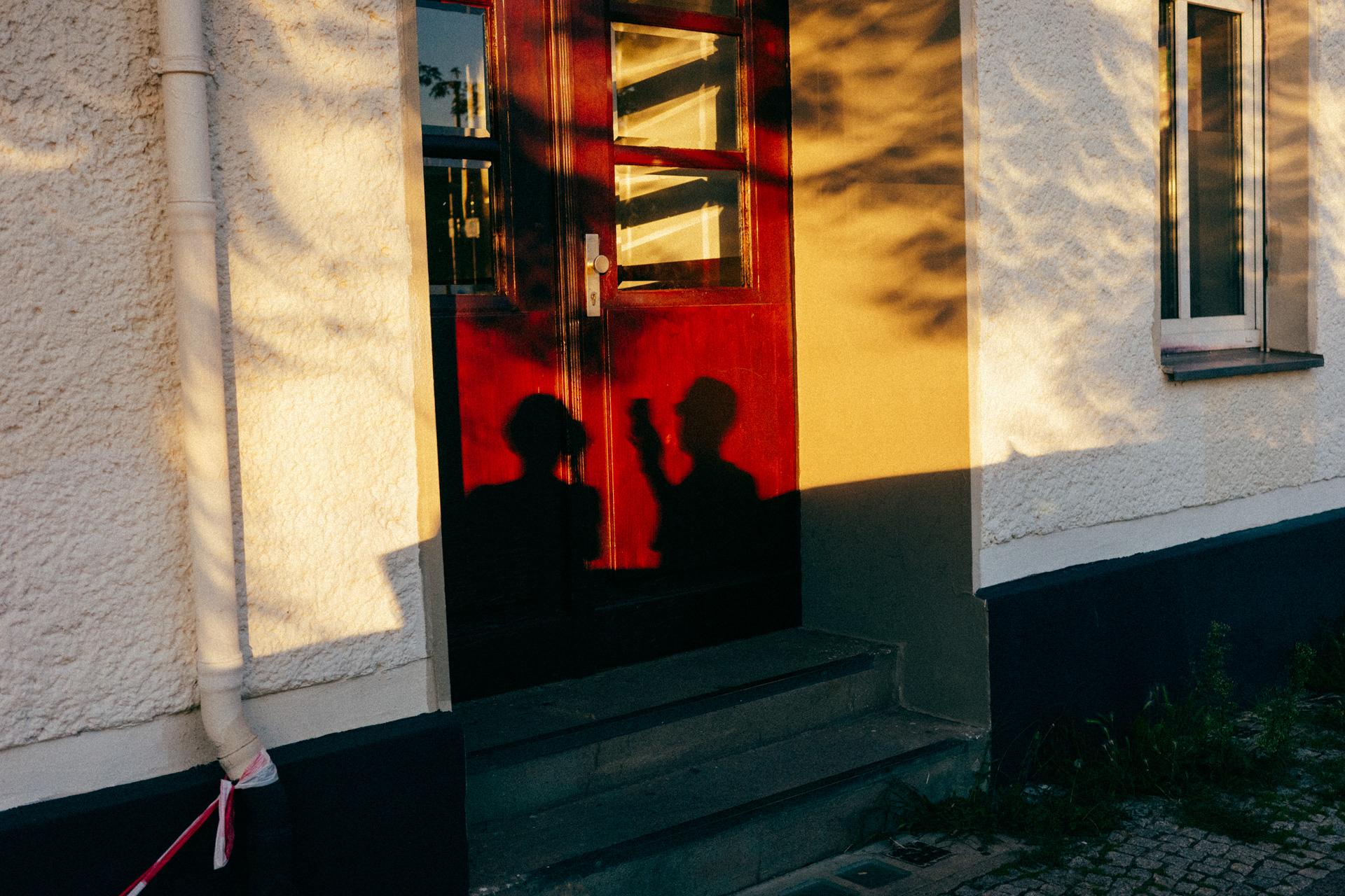 La sombra de dos personas y las sombras de los árboles, con forma de luna, proyectadas sobre la fachada de una casa.
The shadow of two people and the moon-shaped shadows of trees cast onto the façade of a house.