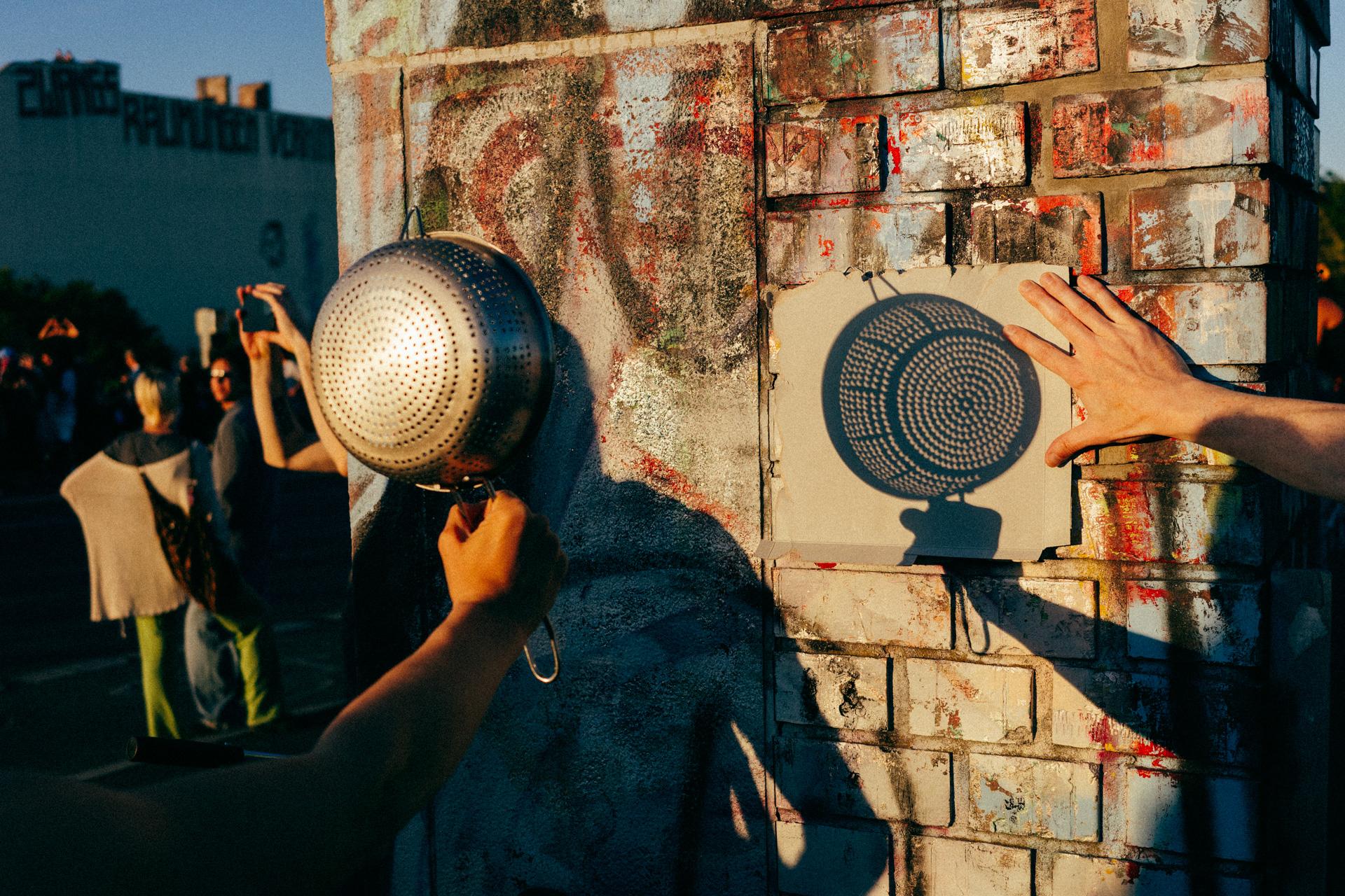 Dos personas proyectando la sombra de un colador sobre una cartulina en un muro de Bösebrücke. Las sombras tienen forma de luna.
Two people casting the shadow of a colander onto a piece of cardstock on a wall at the Bösebrücke. The shadows are crescent-shaped.