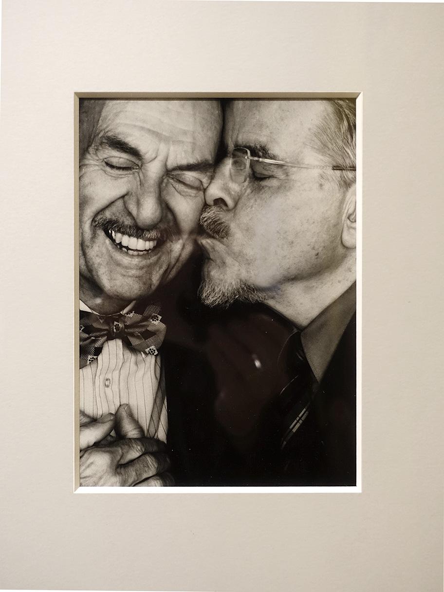 Een foto uit een serie die is gemaakt door Erwin Olaf voor het zestigste verjaardag van het COC, de oudste nog bestaande belangenorganisatie voor lhbti+'ers ter wereld. Een portret van twee wat oudere mannen. Krap uitgesneden qua beeld. De rechter man met bril en gesloten ogen kust hartstochtelijk de linker man op zijn wang die een enorme glimlach heeft door deze liefdesuiting. beide mannen hebben een snor. De linker man is heel netjes aangekleed en heeft een strikje aan zijn boord van zijn overhemd. Linksonder zie je nog net dat de heren elkaar's handen vasthouden.
