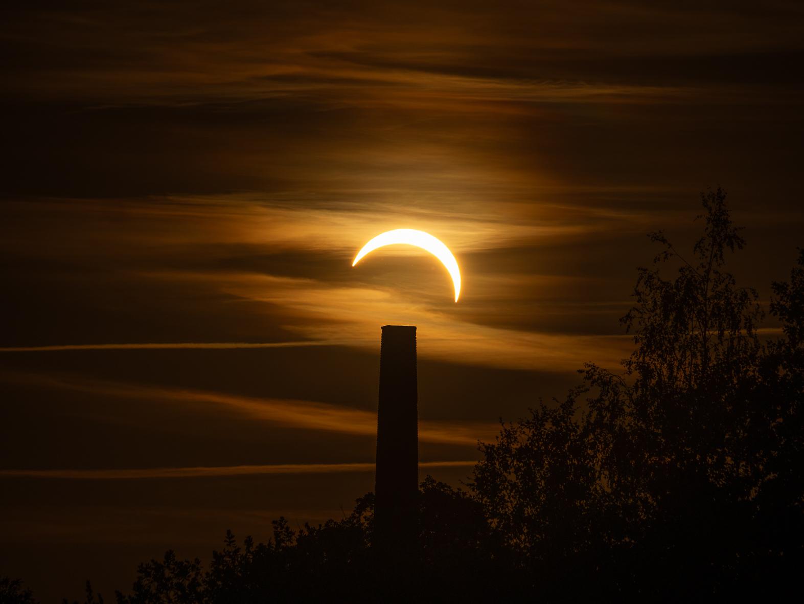 Ein orangener mit leichten Wolken durchzogener Himmel. Im zentrum die nach links unten geöffnete schmale Sichel der Sonnenscheibe direkt über dem Schattenriss eines Alten Schornsteins, der zwischen Bäumen in den Himmel ragt