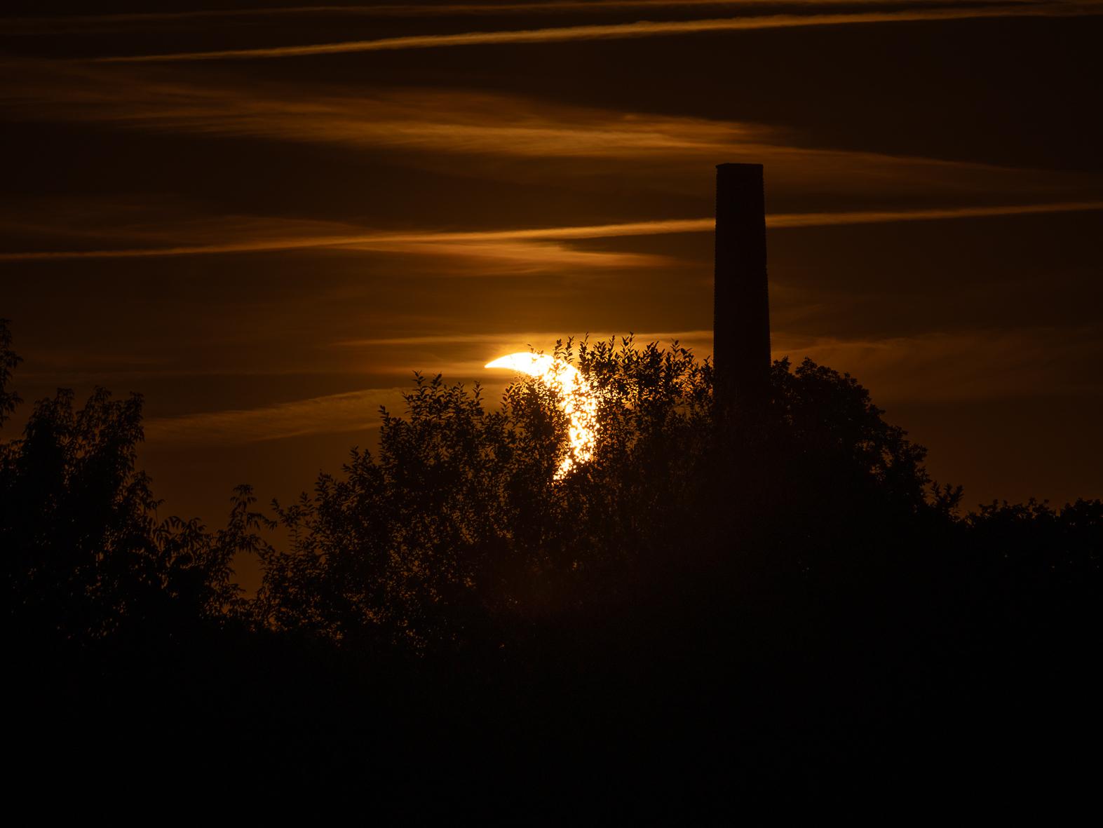 Ein orangener mit leichten Wolken durchzogener Himmel. Links die nach links  geöffnete  Sichel der Sonnenscheibe, rechts daneben der Schattenriss eines slten Schornsteins, der zwischen Bäumen in den Himmel ragt. Die Sonne neigt sich dem Horizont zu