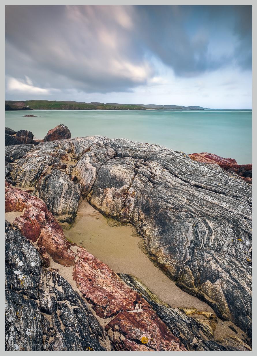 A photograph of Uig Bay on the Isle of Lewis in the Outer Hebrides of Scotland at low tide, showing a rugged shoreline covered with weathered, richly textured rocks, shallow pools of water and areas of pale sand. The rocky foreground leads towards a calm turquoise sea, with the distant coastline beneath a dark, brooding sky.