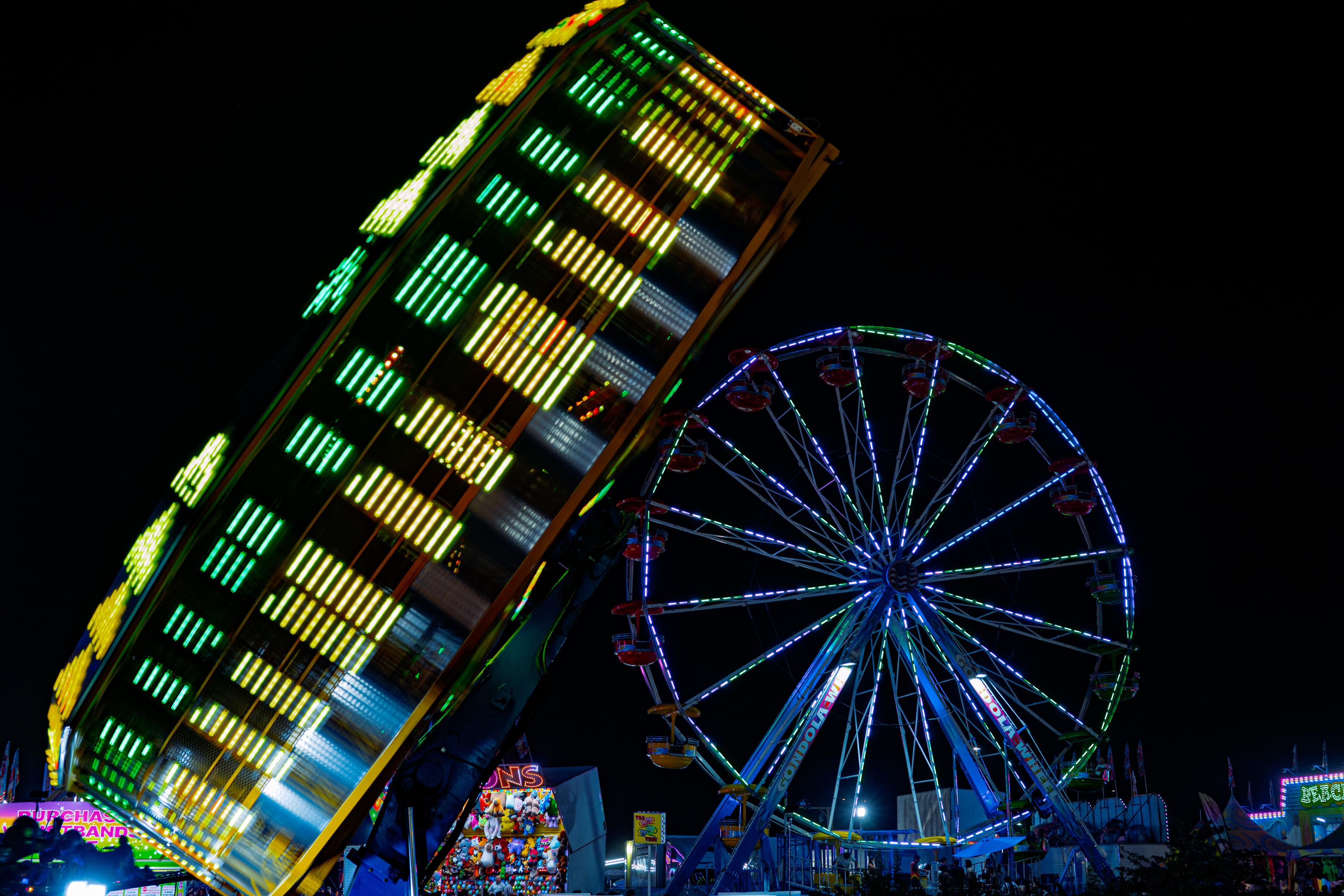A night long-exposure photo at a fair. On the left, a large spinning ride is blurred into vertical streaks of green, yellow, and white light from its motion. On the right, a stationary Ferris wheel labeled “Gondola Wheel” glows with blue and green lights against a black sky. Game booths and other rides are lit up along the bottom of the frame.