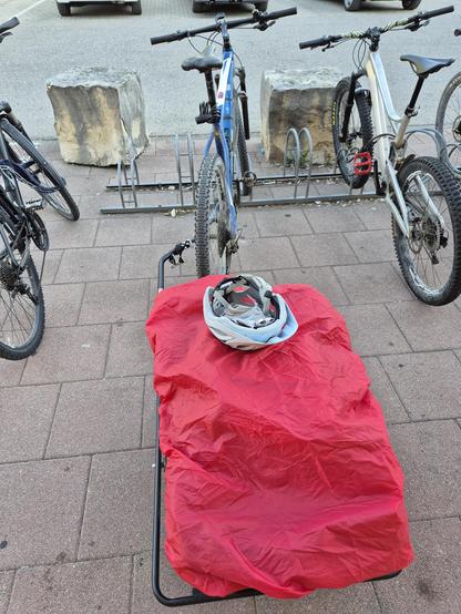 This image captures a practical and everyday scene at a bicycle parking area. In the foreground, there is a red fabric cover draped over a metal frame, likely used to protect a bicycle or gear from the elements. On top of the cover, a bicycle helmet is placed, suggesting that the owner might be nearby or has temporarily stored their gear.
Behind the red cover, there are three bicycles parked in a bike rack. The bicycles vary in style and color:
- The first bicycle on the left is dark-colored, p…