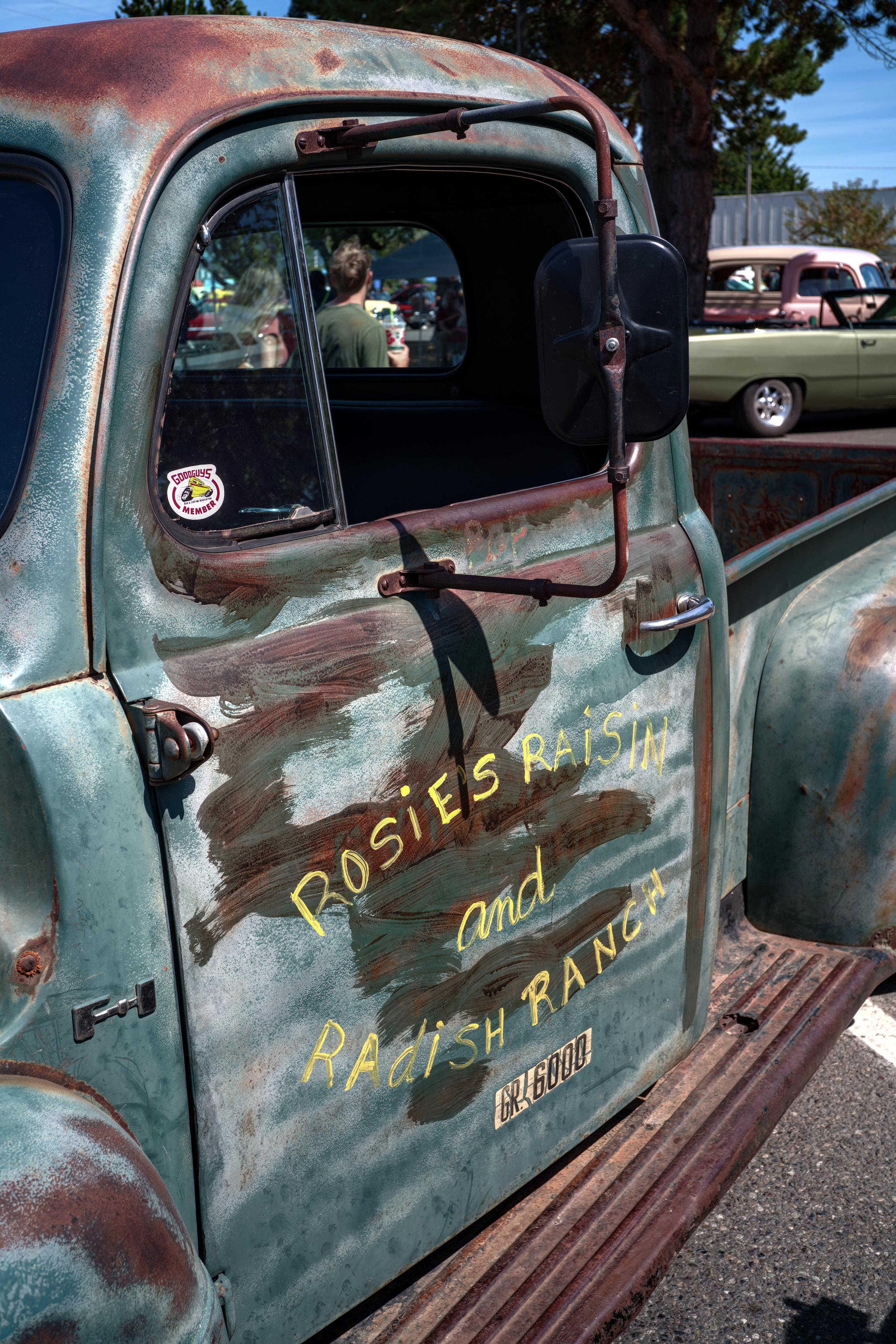 Color photo of a Ford Series F-1 ½ ton truck (model years 1948–1952) in portrait format, taken of the driver's side door.

Unlike most classic cars you see at a vintage car show, which are spotless and shine line a well polished mirror, this F-1 shows its age and is  heavily oxidized: the sidestep in its entirety; the roof, driver side door, front and rear fender flares in large swaths, and the bracketed side view mirror in its entirety except for the black painted backside of the mirror itself. The truck's base color paint is a light turquoise shade.

The driver's side door has a horizontally centered block of text in light yellow:

Rosies Raisin
        and
Radish Ranch

And is swathed with a rust colored finish with a hand brush, painted horizontally across the door.

Underneath that block of text is another; black lettered "GR. 6000", with an off-white stenciled outline.

In the background: a couple walking away from the camera through the rear cabin window; an olive green converti