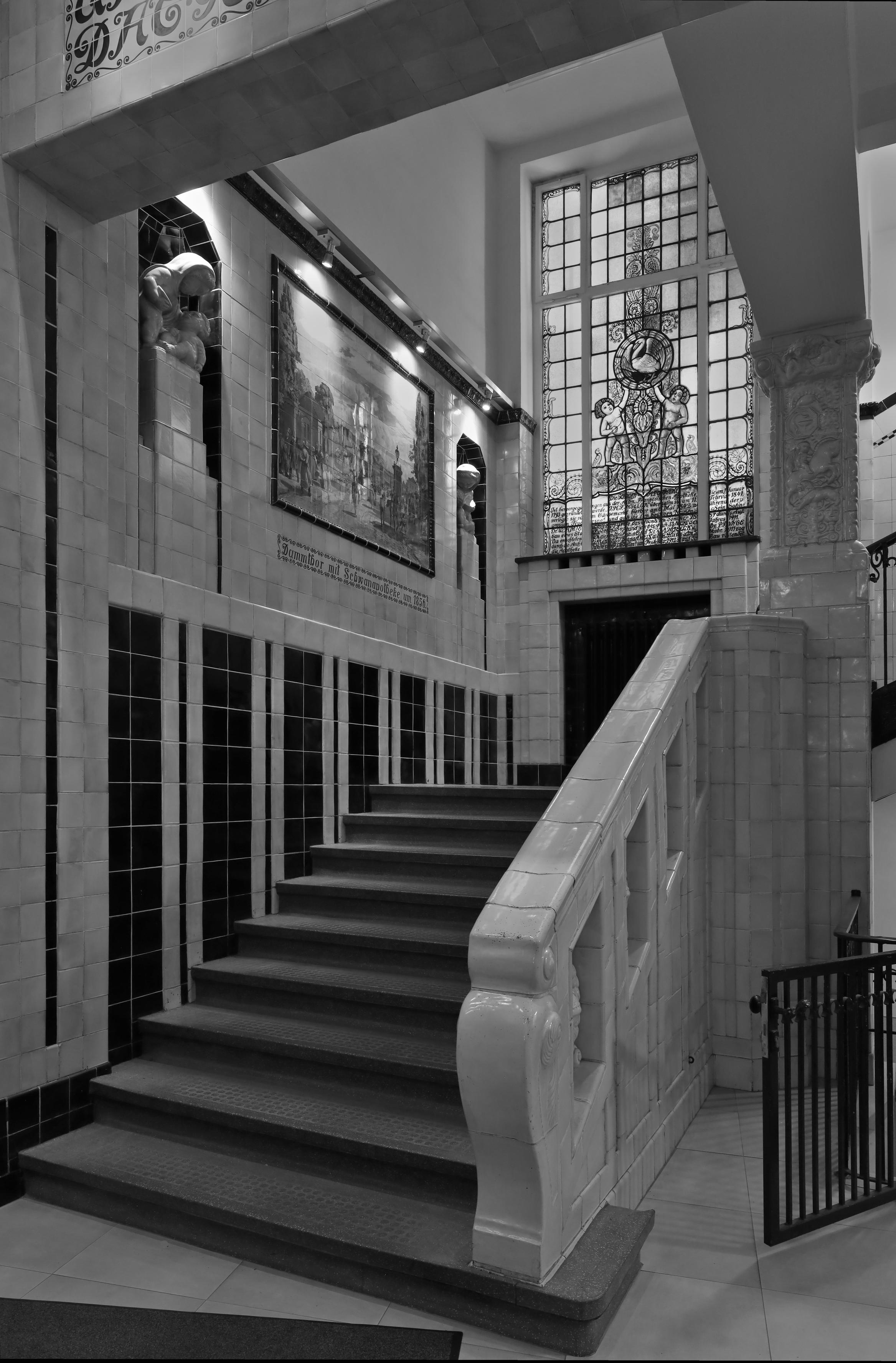 Schwarzweiss-Foto des unteren Einstiegs in ein historisches Treppenhaus in Hamburg mit Fliesen an den Wänden und einem keramischen Handlauf. Das Außenfenster rechts oben im Bild besteht aus vielen bunten Elementen und stellt ein Bild dar.