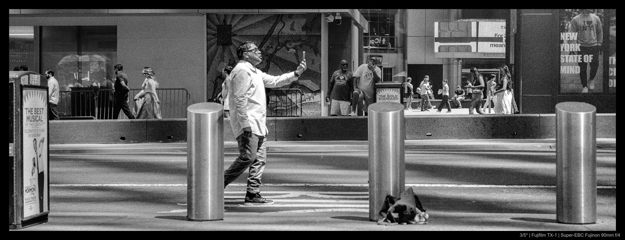 A person raises their phone in front of them to snap a selfie while walking down the side of a street in Times Square, standing to the left of the protective bollards.