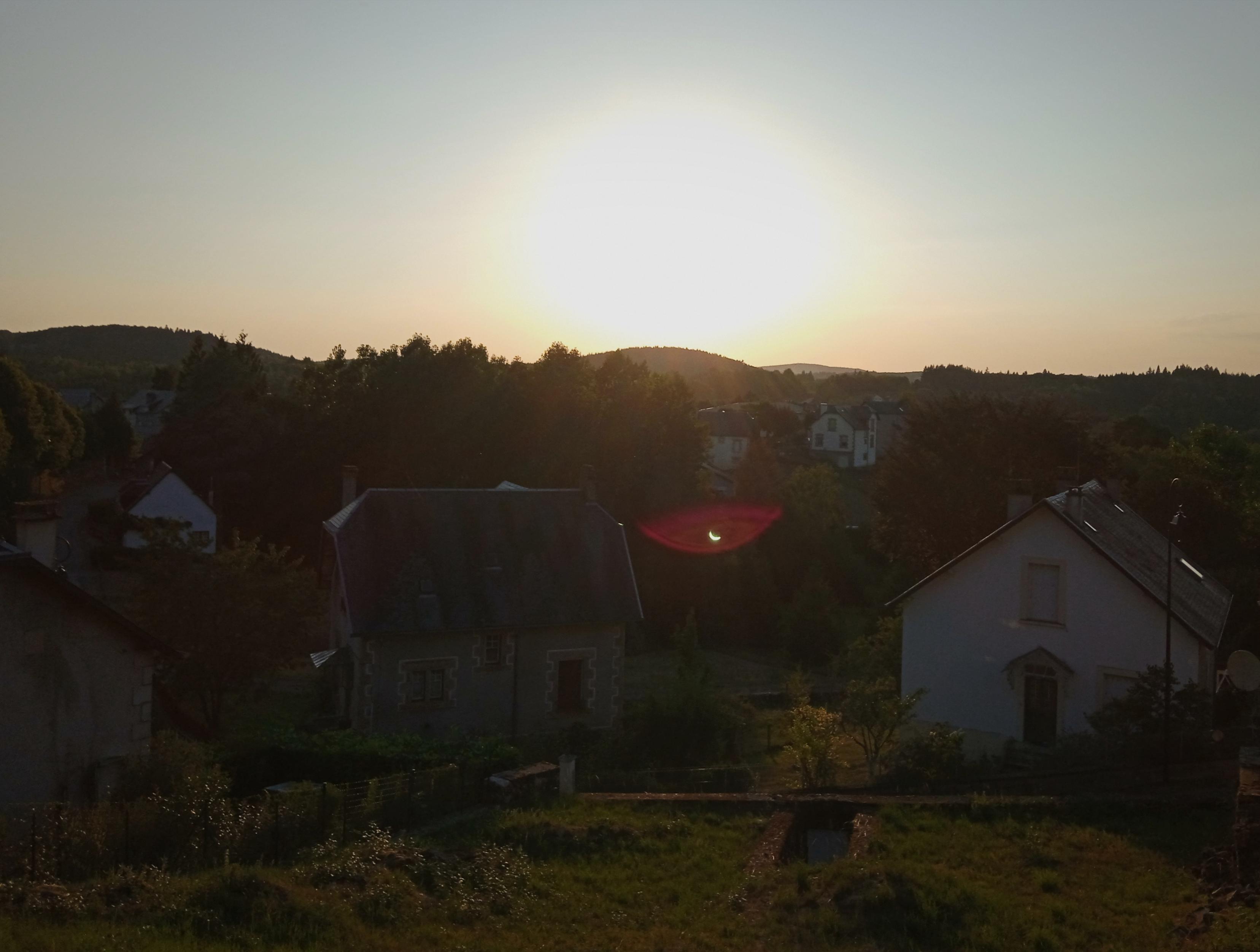 Dark village and trees, under an over-exposed orange white sun low in the sky. The lens flare has a tiny crescent artifact - the reversed, partially eclipsed sun!