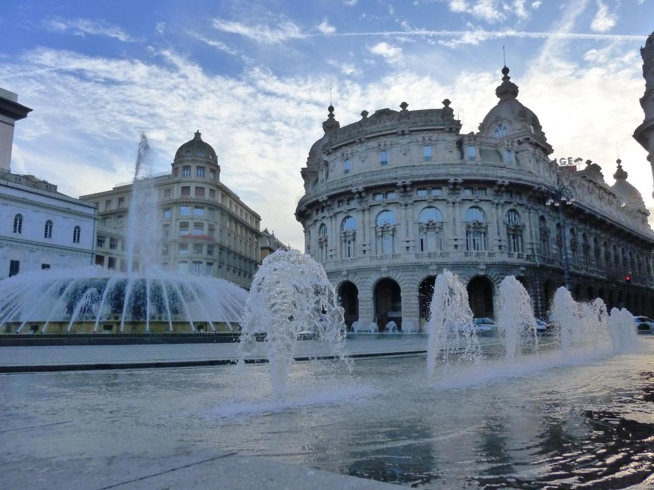 Das Bild zeigt einen sehr großen, kreisförmigen Springbrunnen. In seiner Mitte steigt ein Wassersrtahl auf umgeben von Strahlen, die von außen im Bogen zur Mitte spritzen.. Im Vordrrgrund rechts sind weitere, kleinere Fontänen zu sehen. Im Hintergrund stehen alte Gebäude mit prachtvollen Fassaden mit Rundbögen.