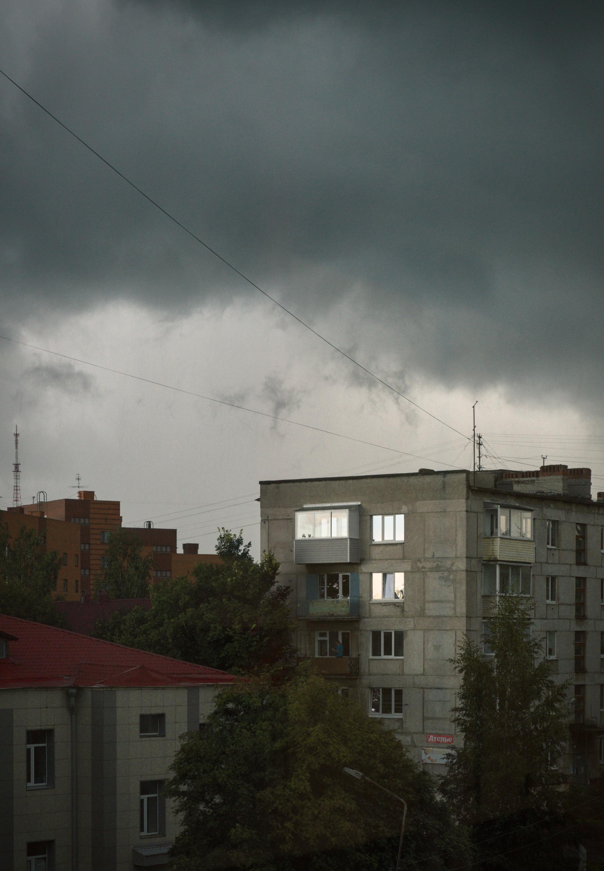 This color photograph captures an urban landscape in the hour before a storm. The upper part of the frame is occupied by a dense, heavy sky covered in dark gray, almost black clouds that loom over the city. Thin dark wires stretch across the sky, adding a graphic quality and a sense of tension to the frame. In the center of the composition is a gray panel apartment building with several enclosed balconies that stand out with their light frames against the gloomy walls. On the left, a building with a red roof and a tall antenna are visible. Below, there are tree canopies and bushes. The image conveys a feeling of calm before the storm, eerie silence, and the harsh beauty of urban weather.