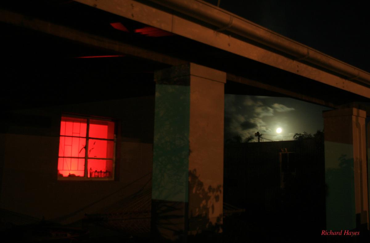 The pillars and part of the roof of a veranda acts as a frame for the moon surrounded by clouds and a window with a red light in theroom behind the window.
The red light tints the white pillars and veranda ceiling
Street l8ghts cast a pale yellow light on the pillars