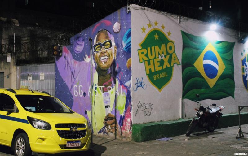 Graffiti in the streets of Lapa, featuring a black man with black glasses smiling on the left wall of a corner.

Bahian singer Léo Santana has been honored by the NegroMuro project (IG: negromuro + _fernandosawaya ), which celebrates prominent figures of Black culture in Rio de Janeiro. This week, a special mural was created to mark the release of his new album, “GG Astral.” Located at 180 Rua do Rezende in the city center, the mural was painted in just one day as part of a promotional campaign.

On the right, a Brazilian flag on white background and a Coat of arms in green with 5 yellow stars above and text in the center “RUMO ao HEXA Brasil”.

Signed by jambeiroarte
Marcos Alexandre Jambeiro