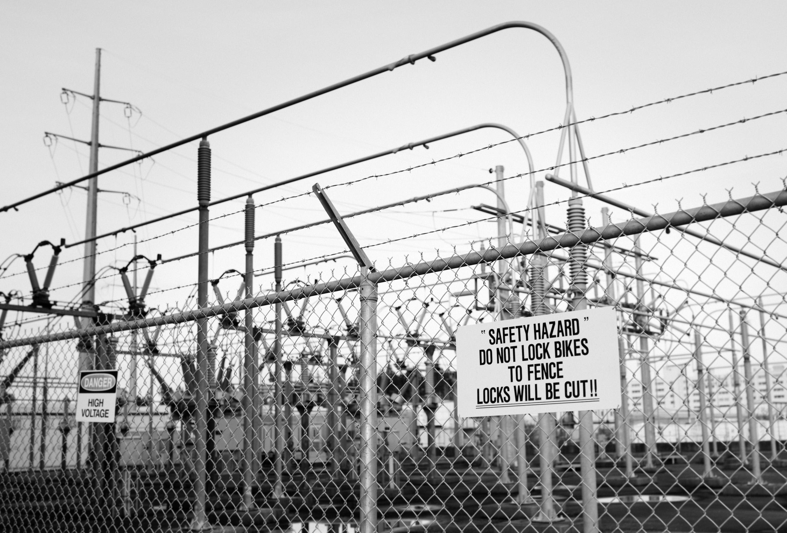 Black and white photo of a power substation in landscape format.

The foreground has a chain link fence running horizontally across the frame from left to right at a 30 degree angle with a vertical support pole in the middle. To the right of the support pole, a white sign with black text horizontally centered reads:

"SAFETY HAZARD"
DO NOT LOCK BIKES TO THE FENCE
LOCKS WILL BE CUT

Behind the fence in the background are various current carrying components.

Obviously people lock their bikes to the fence, which is why the sign was posted. But it still begs the question: how could anyone lack the common sense to not figure this out themself?!?!
