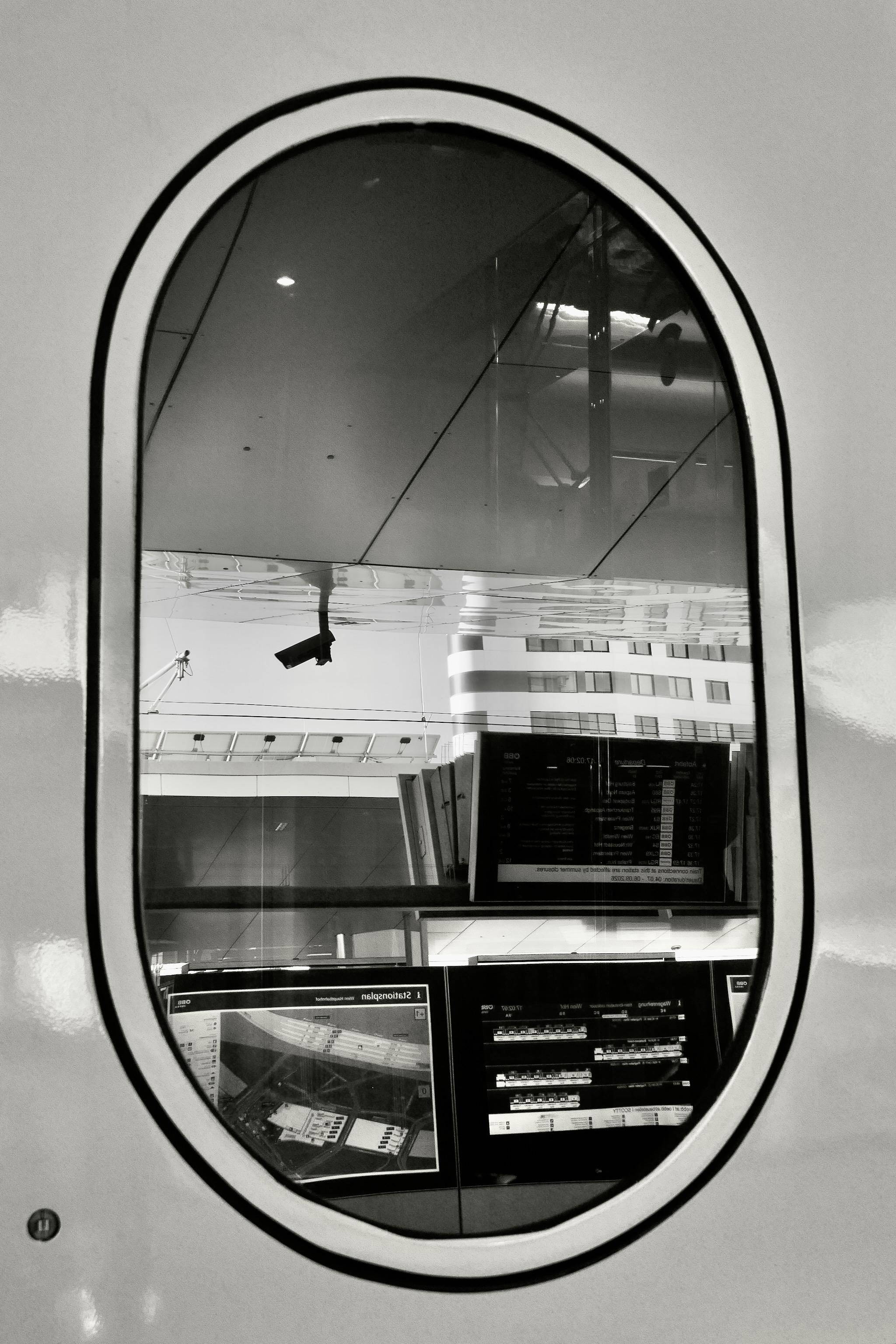 Picture is showing a train window in the door of an ICE train. Its shape is oval, like a window of an airplane. In the window reflections of the platform are visible: survaillance camera, monitors, the roof of the platform and in the background are some multiple-level buildings. Picture is black and white and was edited to brighten the shadows and dark areas to make details visible.