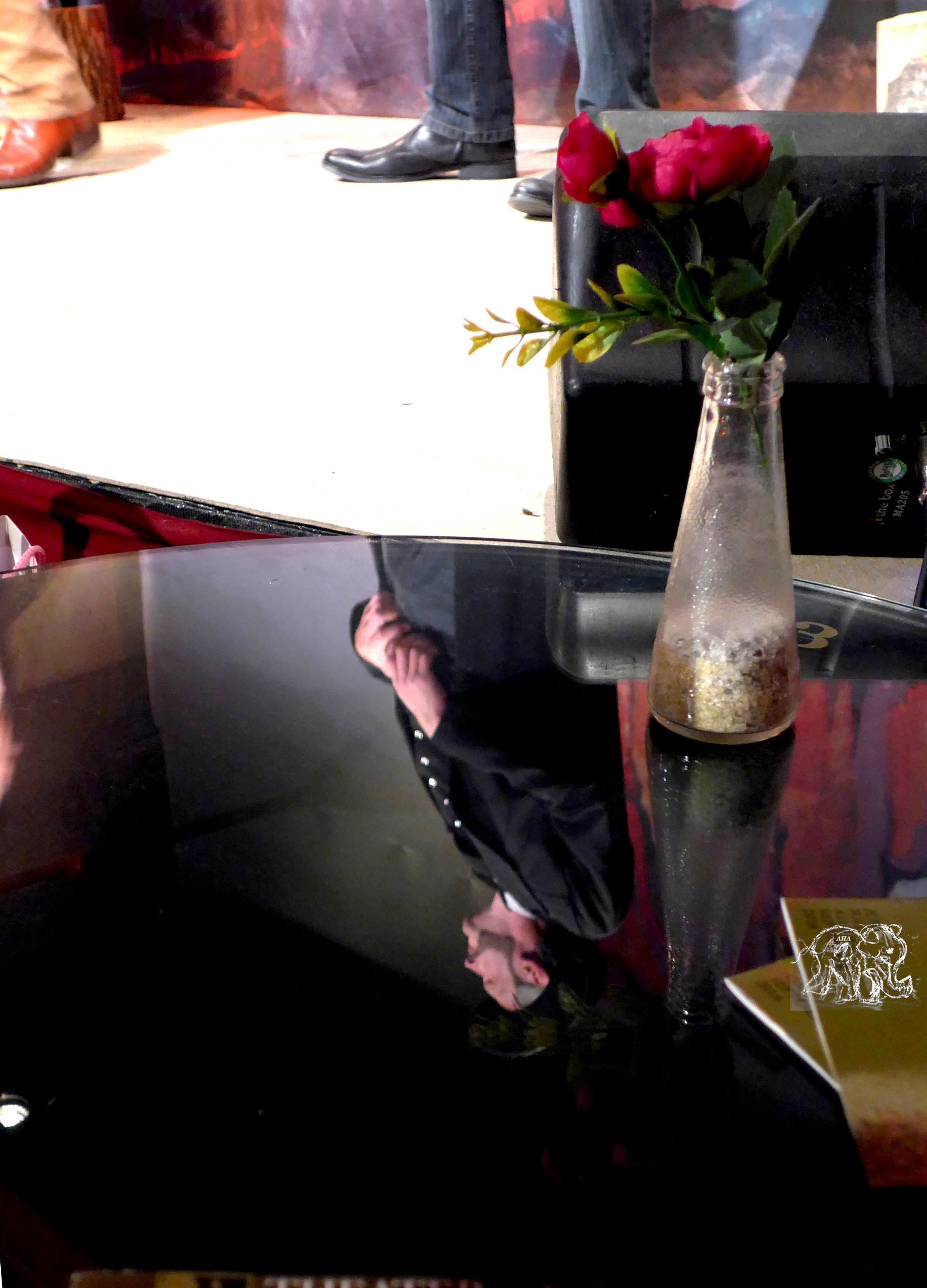 A black table in front reflects a stage scene. On the table there  is a vase with roses and behind you see the bright stage and 3 feet belonging to  actors the right one covered by a dark loudspeaker.
