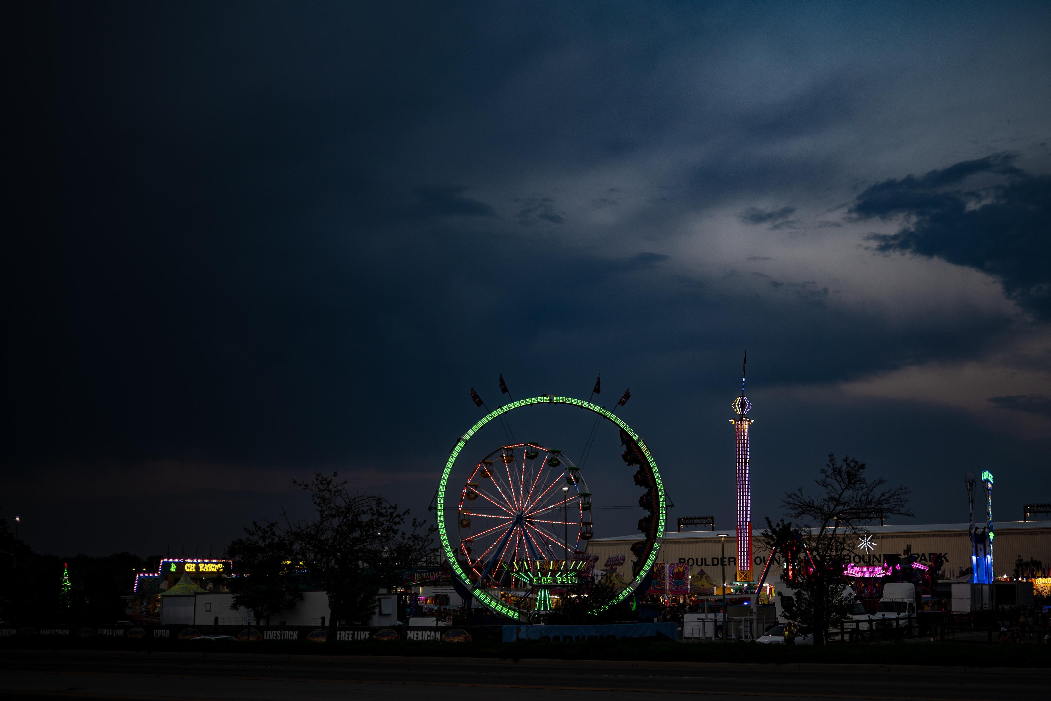 A wide blue-hour view of the Boulder County Fair under a large, dark, cloudy sky. In the center, a looping roller coaster lit in green light encircles a Ferris wheel glowing red and white behind it. A lit drop-tower ride stands to the right, with more rides, game booths, and the fairgrounds building spread across the horizon.