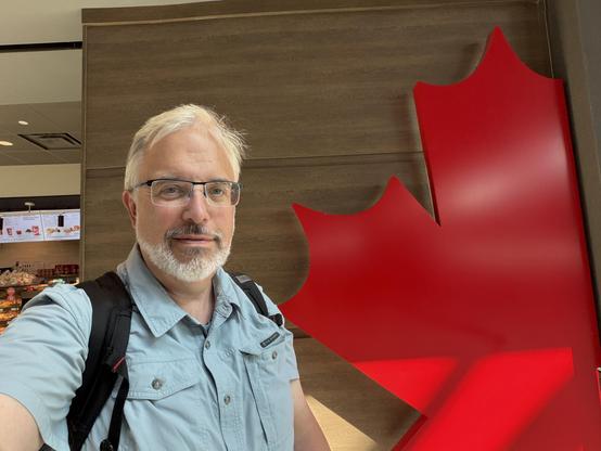 A man with glasses and a beard poses for a self-portrait in front of a large red maple leaf design in an indoor setting, possibly a café or airport. He is wearing a light blue shirt and has a backpack.
