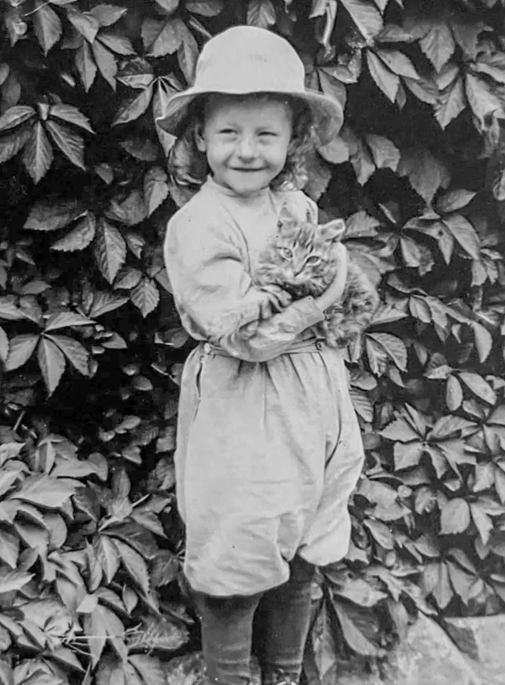 black and white photo of a light skinned child standing in front of a wall of foliage, smiling and holding a longhaired tabby kitten in their arms.