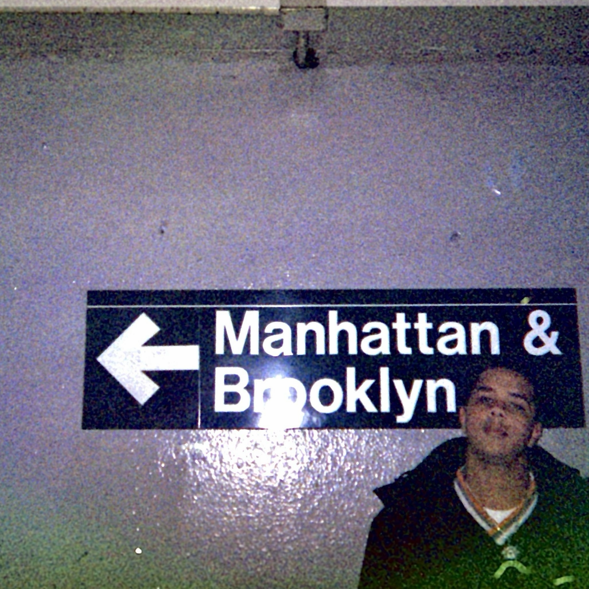Greeney photo of me, bottom right, in a big coat. I am standing in front of a subway sign that says Manhattan and Brooklyn with an arrow pointing left. Behind me is a cement wall. A green tinge covers the bottom of the photo because it is old