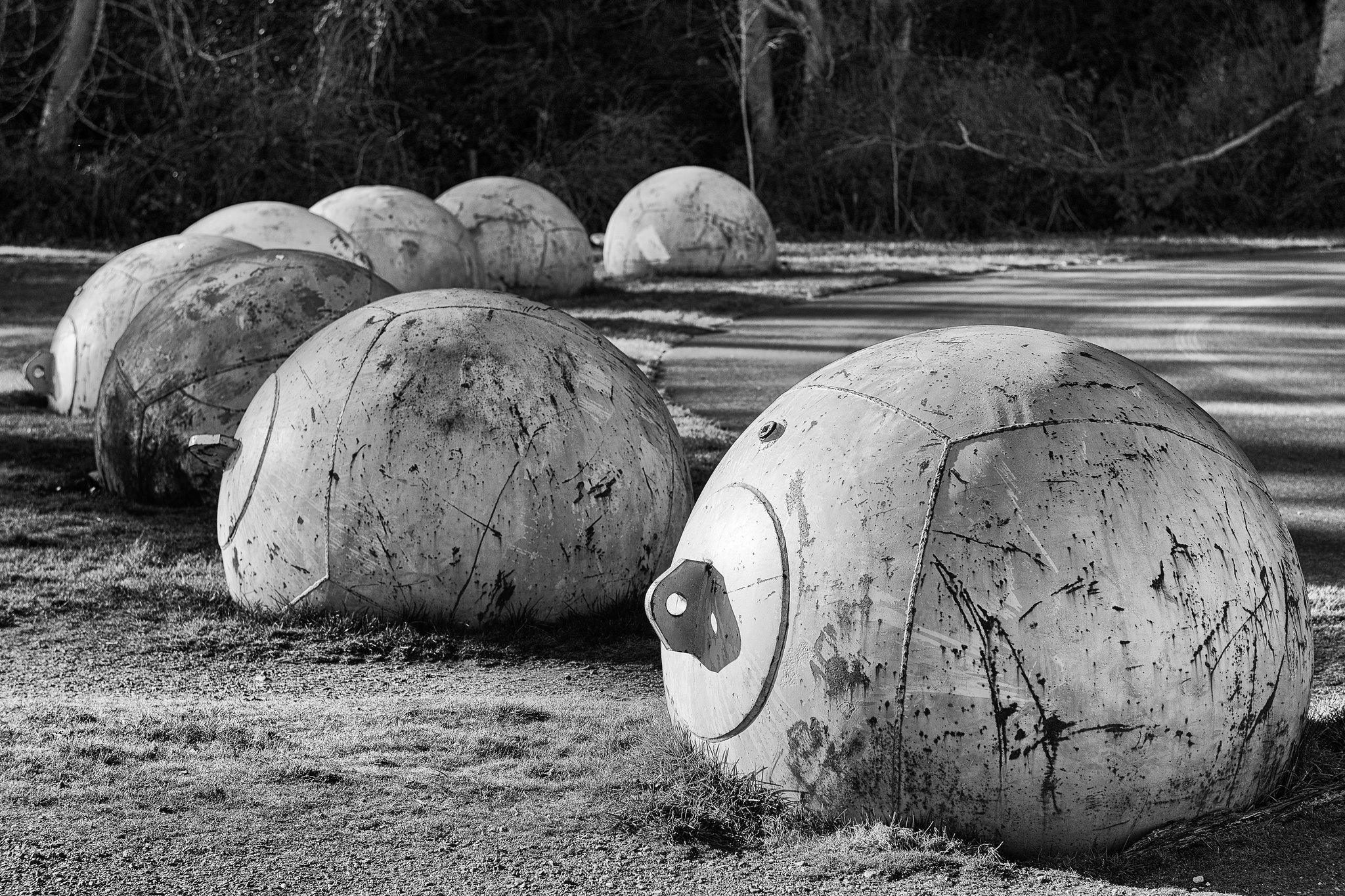 Black and white photo in landscape format of eight empty (disarmed) naval sea mines arranged in a half circle 1-2 meters apart along the outside of a curved roadway. Each mine is buried about a quarter of its total size. 

Long late afternoon shadows from the mines and trees in the background cover the road in approximately at a 4 o'clock angle.