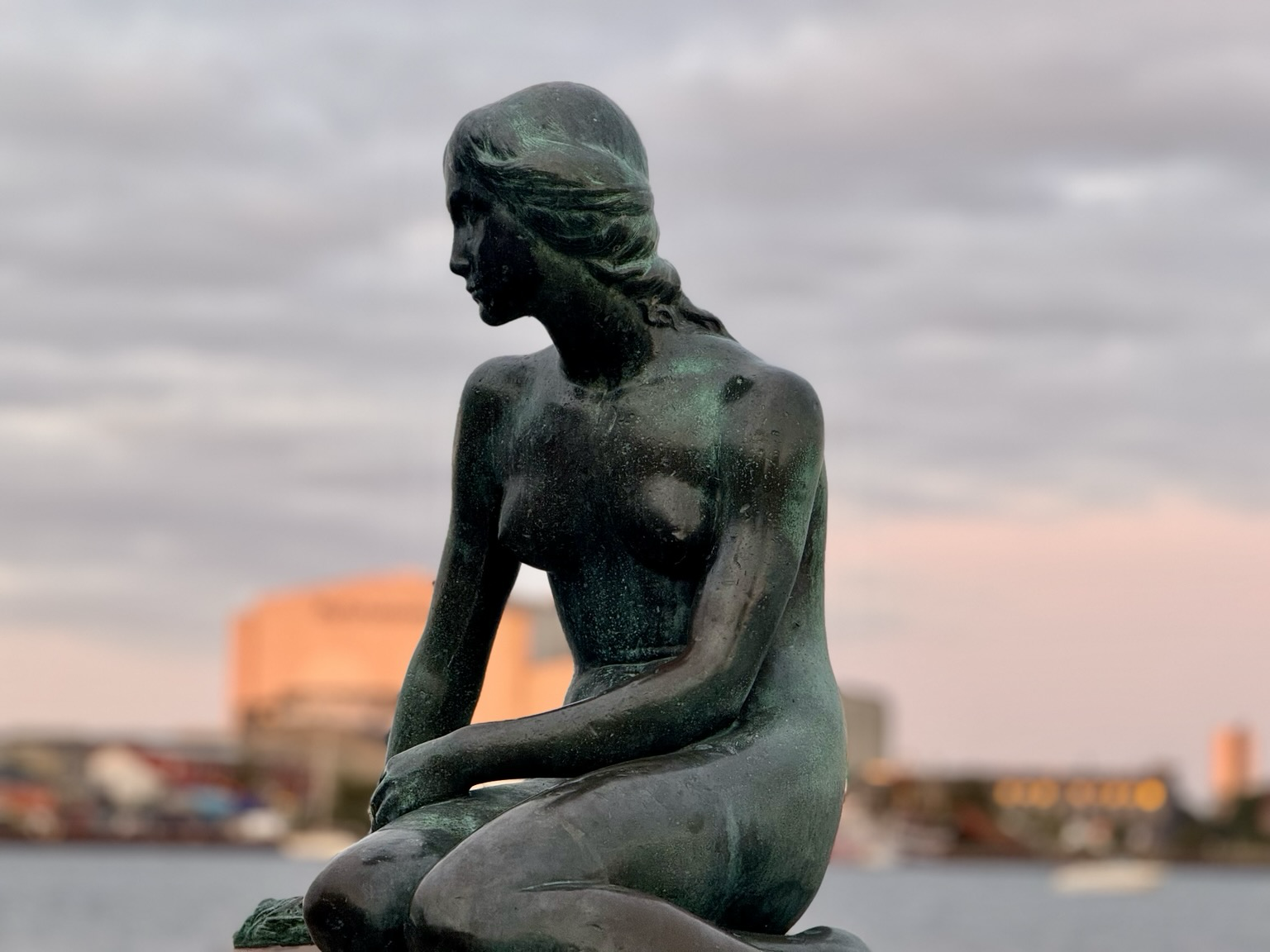 The bronze statue of The Little Mermaid in Copenhagen, sitting in profile by the water. A cloudy sky and softly blurred buildings lit by warm evening light can be seen in the background.