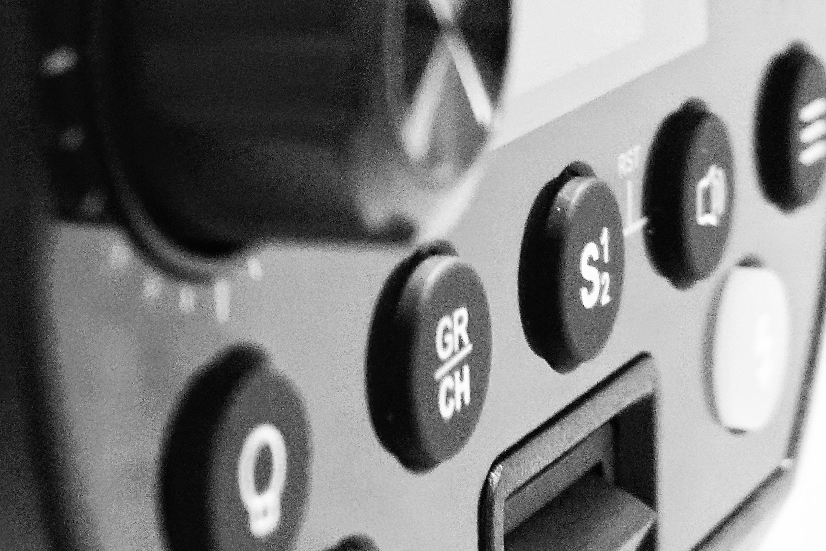 Zbliżenie w skali makro przedstawiające panel kontrolny profesjonalnej lampy studyjnej (Studio Stawki) w kontrastowej, czarno-białej tonacji. W ostrości znajdują się fizyczne, gumowane przyciski sterujące z wyraźnymi oznaczeniami funkcji, takimi jak zmiana grupy i kanału (GR/CH), tryby fotoceli (S1/S2) oraz sterowanie światłem modelującym. W tle widoczny jest fragment obudowy oraz wyświetlacz LCD.
