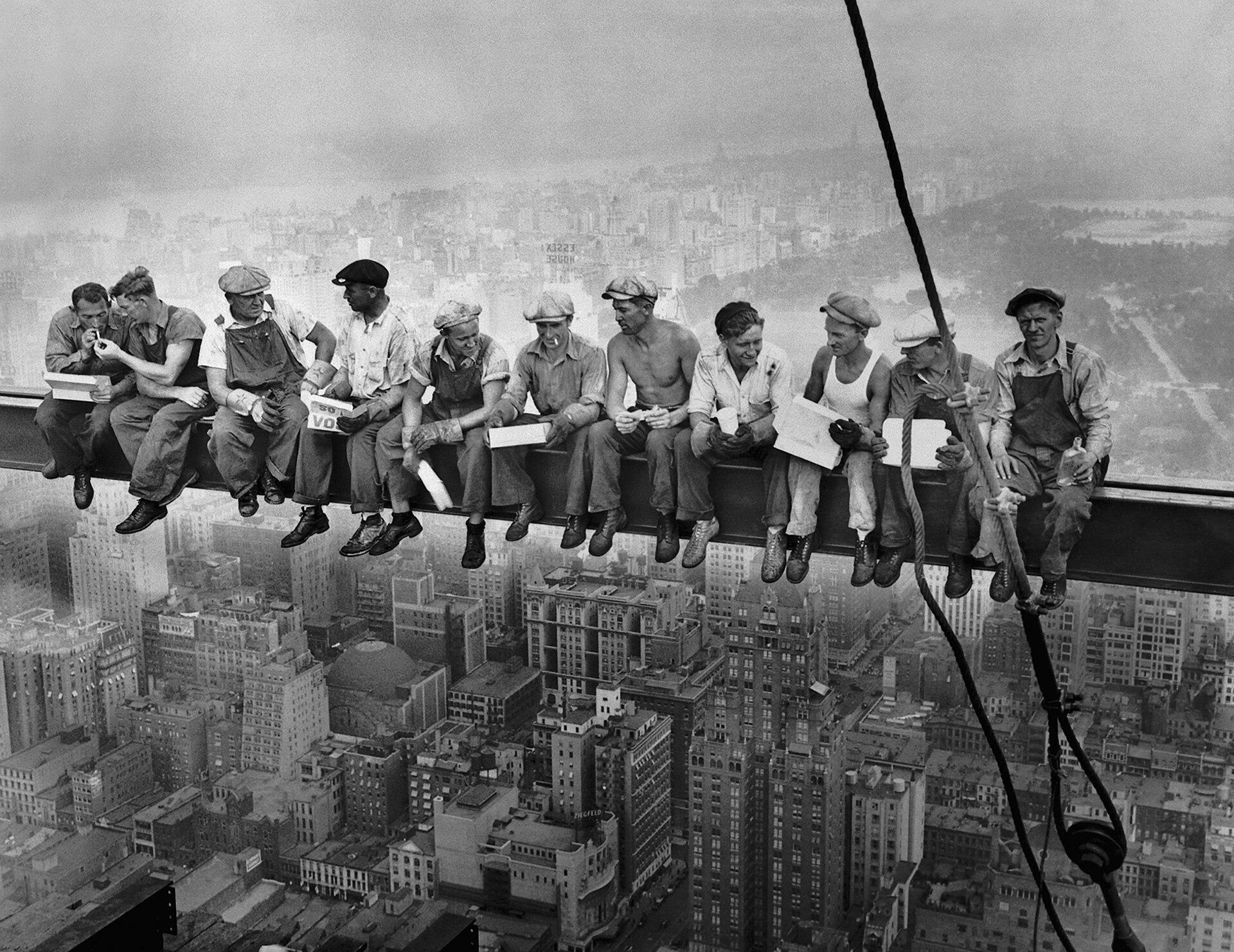 Fotografia in bianco e nero. Una fila di operai edili siede su una trave d’acciaio sospesa a grande altezza sopra Manhattan. Gli uomini, vestiti con abiti da lavoro e berretti, mangiano, fumano e conversano mentre tengono i piedi penzoloni nel vuoto. Sullo sfondo si estende il panorama urbano di New York, avvolto dalla foschia, con numerosi grattacieli e edifici sottostanti.
