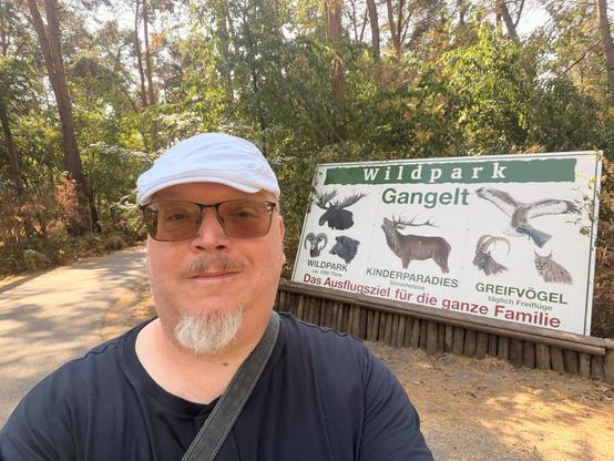 Mann in dunkler Kleidung posiert vor einem Schild mit Tierbildern in einem Waldgebiet.