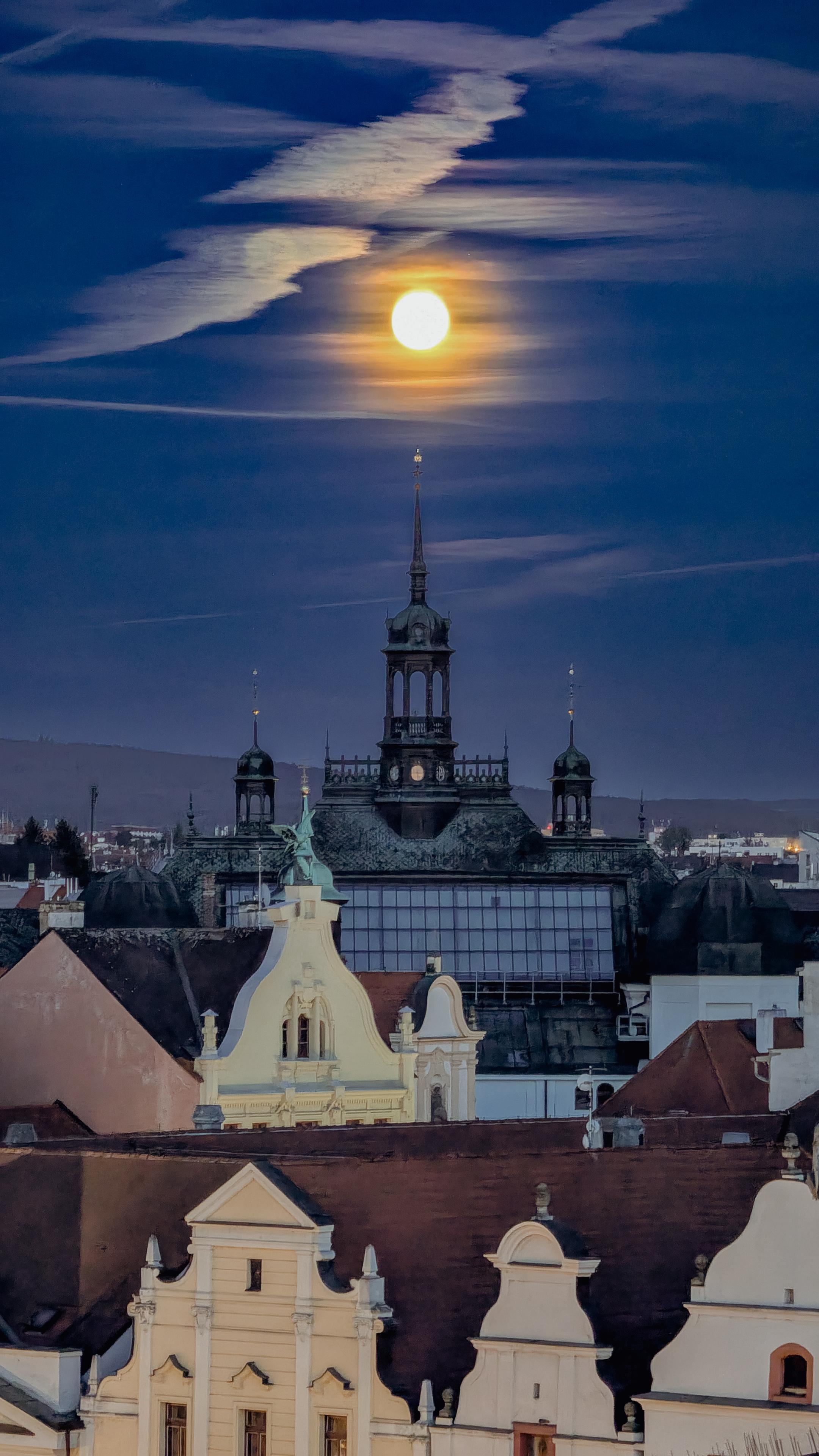 ​A night view looking over the decorative rooftops and spires of buildings in the historic center of Plzeň. A bright, large full moon is high in a blue sky above the main central tower, illuminating long, wispy clouds.