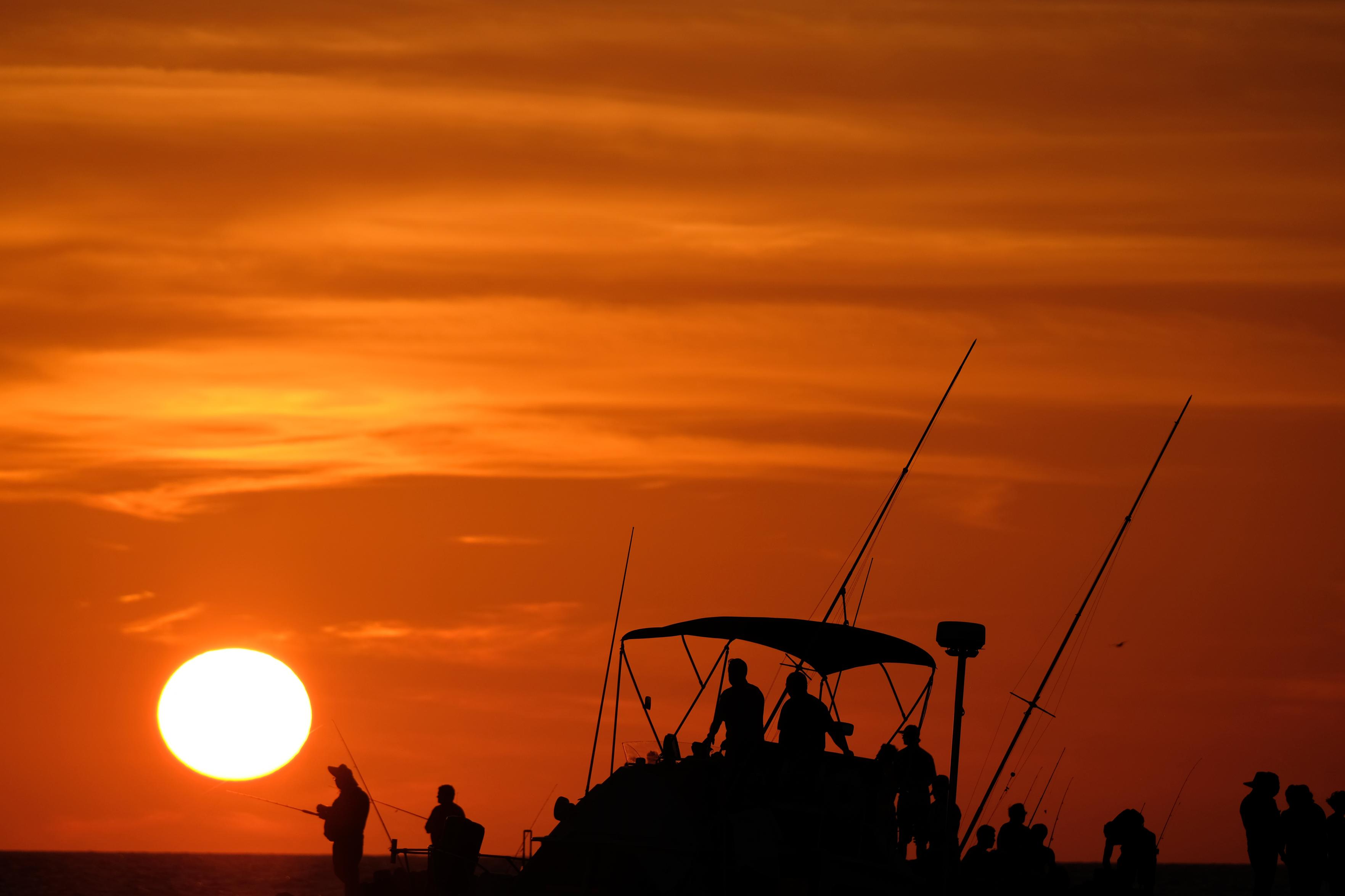Photo by Fenichel

Most of the frame is a vivid orange sunset, streaked with white/orange clouds, and the setting sun in lower left, at horizon (sea level).

In the center is the silhouette of a fishing boat coming in for the day, passing by a jetty with several fishermen also in silhouette against the intense sunset, behind and above them.