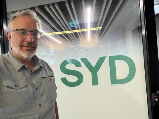 A person with gray hair and a beard stands next to a glass door featuring the large letters "SYD." The background shows a modern indoor space with minimalistic design and indirect lighting.