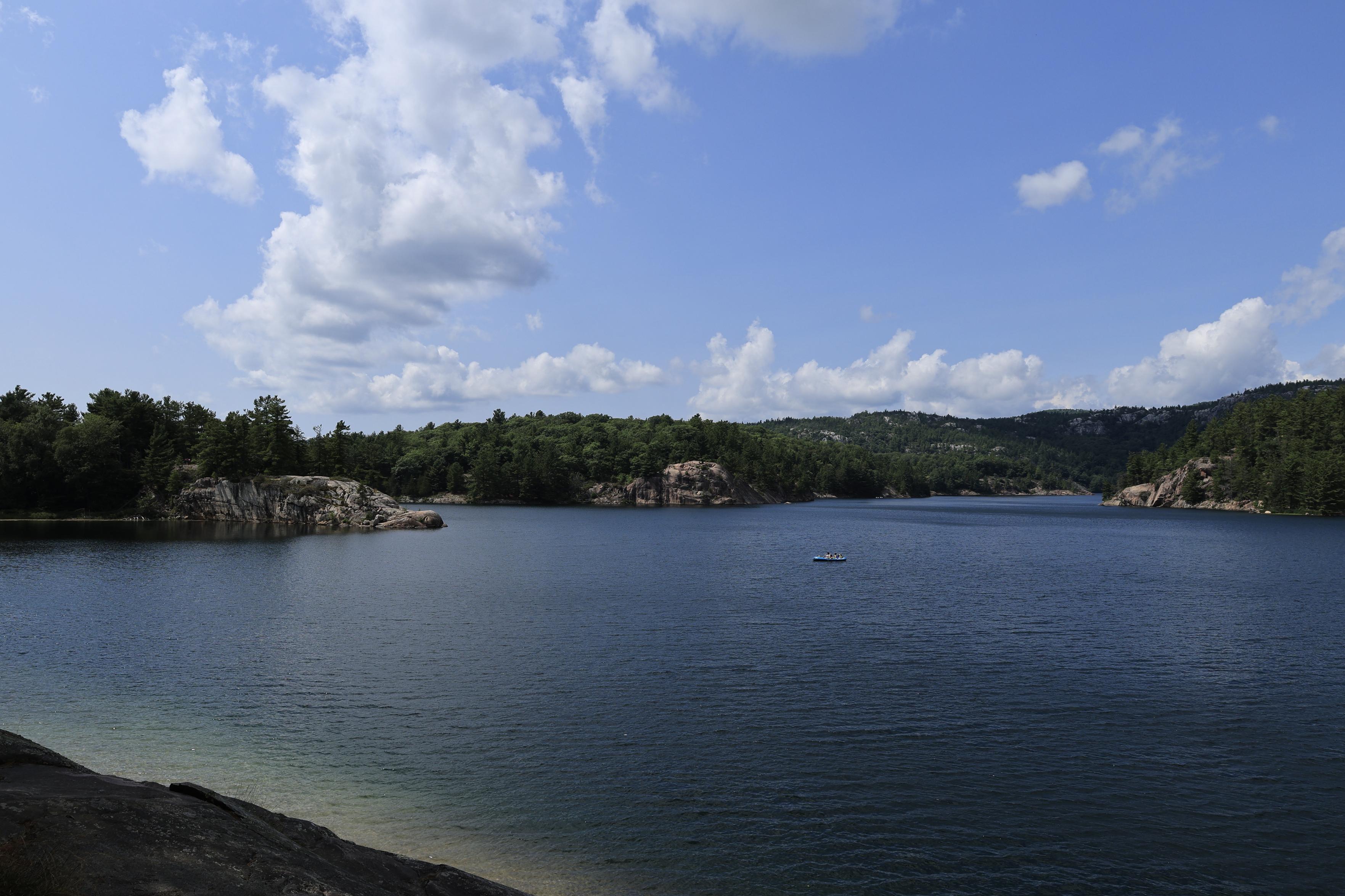 This is a landscape photo taken in the summer from an elevated viewing point  looking out over a smaller inland lake.  The water is a vivid blue which contrasts with the green of the forested areas around the shoreline. Various rock formations can also be seen around the shoreline. Some white clouds are present in the sky overhead.