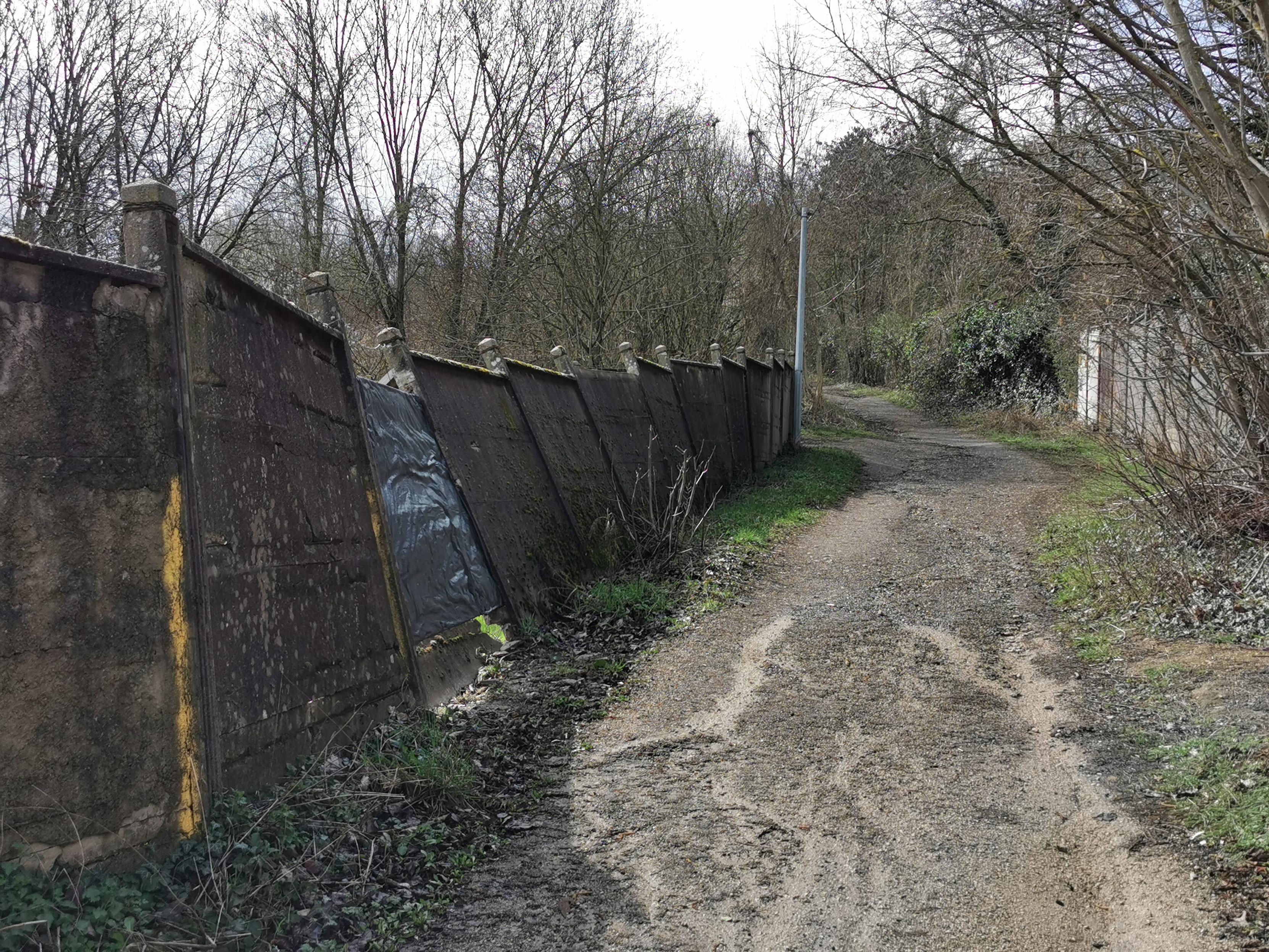 Chemin longé par un mur en éléments de béton préfabriqués partiellement effondrés.