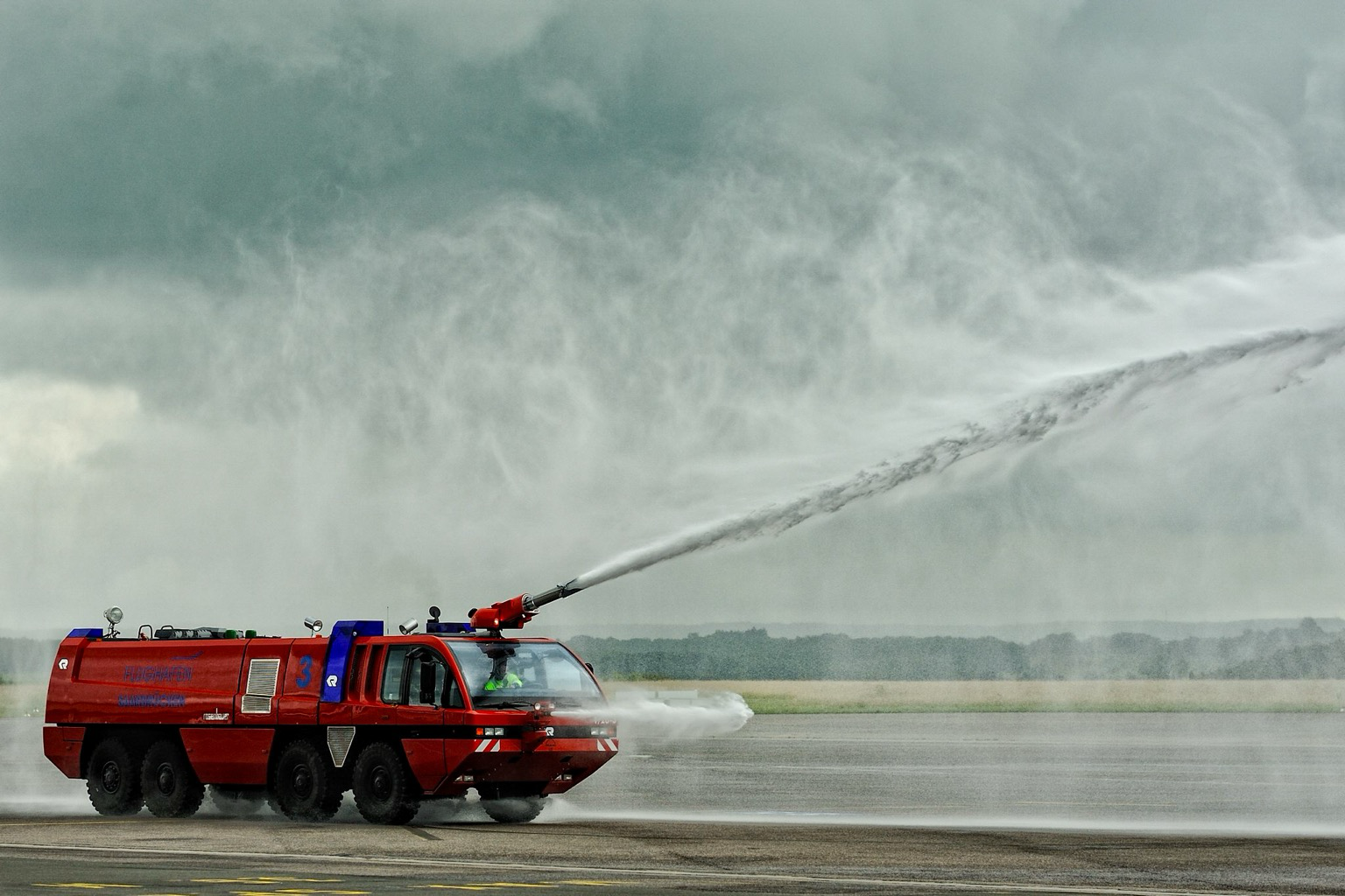 Bildbeschreibung (Deutsch)  * Zusammenfassung: Ein rotes Flughafenlöschfahrzeug sprüht Wasser auf einem Flugfeld.  * Vordergrund: Ein großes, achträdriges, rotes Flughafenlöschfahrzeug bewegt sich auf einer asphaltierten Landebahn. Ein starker Wasserstrahl wird hoch und weit von einem Dachmonitor nach vorne-rechts gesprüht, der als Gischt in der Luft sichtbar ist. Ein weiterer, kleinerer Wasserstrahl spritzt von einer Stoßstangendüse nach vorne. Die gläserne Fahrerkabine an der Front zeigt eine Person mit einer gelben Sicherheitsweste am Lenkrad. An der Seite des Fahrzeugs sind Aufschriften sichtbar, darunter "FLUGHAFEN..." (der Rest ist unleserlich) und die Nummer "3". Auf dem Asphalt links sind gelbe Markierungen zu sehen.  * Mittelgrund: Eine offene Fläche eines Flugfeldes, gefolgt von einer grasbewachsenen Fläche mit einer Baumlinie in der Ferne.  * Hintergrund: Der Himmel ist vollständig mit grauen, schweren Wolken bedeckt. Image Description (English)  * Summary: A red airport res