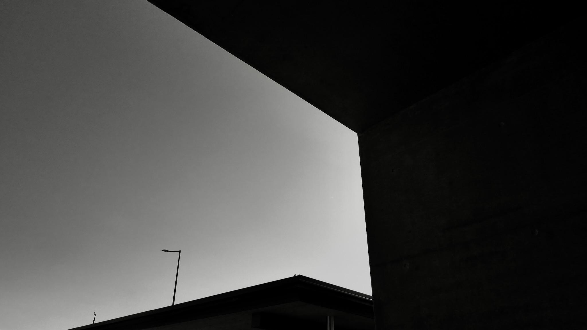 Black and white picture of geometric forms. on the right side is big area of dark black. In the middle on the bottom is a triangle roof with a street lantern in the background. The left hand side is open to the sky in grey. The sun seems to be hidden on the right side in a corner of the shapes.