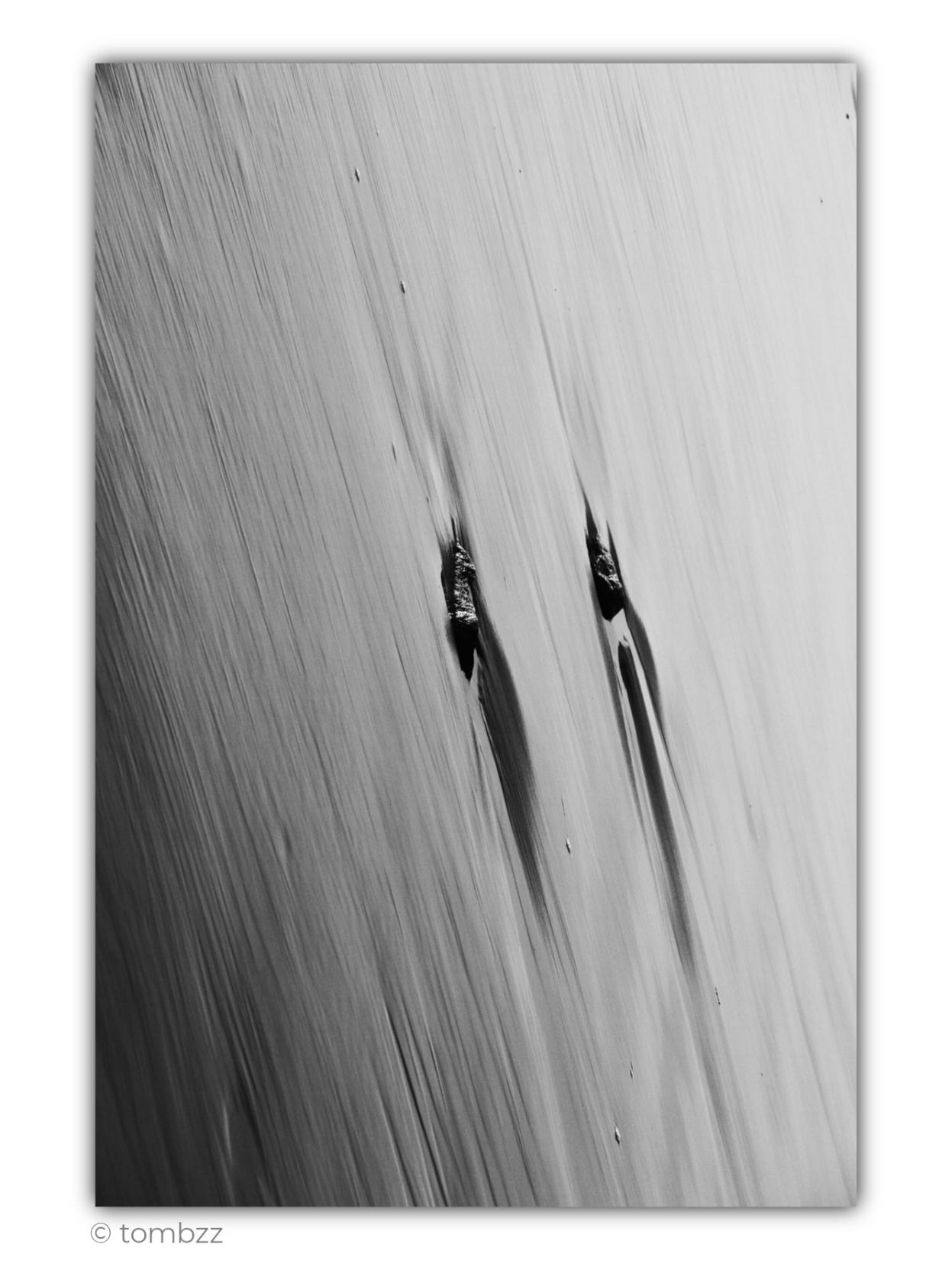 Black-and-white photograph showing wet sand along the shoreline, moments after a wave has receded. The image is framed at an unusual diagonal angle, making this familiar fragment of the beach lose its obvious character and take on an abstract appearance. Fine, parallel grooves left by the water run across almost the entire frame, creating a delicate, fibrous texture. Near the centre, two small dark stones are partially embedded in the sand. Behind each one stretches a narrow channel carved by the retreating water, followed by an elongated shadow. The surface of the sand gradually shifts from deep grey on the left to almost white on the right, creating a subtle transition of light and shade. The whole image resembles an abstract drawing or the texture of a material more than a traditional seaside landscape.