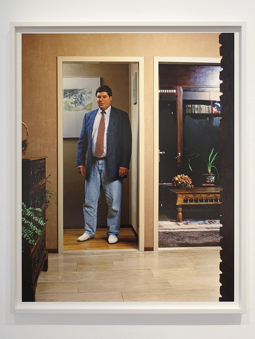 Een kleurenfoto vanuit de hal waar tegenover in een deuropening een volwassen man staat in een blauwe spijkerbroek en een blauw colbert en een rood gemêleerde stropdas op een wit hemd. De houding en de aankleding werkt vervreemdend in deze onnatuurlijke scene. Aan de rechterzijde is nog een deuropening met nog vaag een antiek houten salontafel en daarachter een zwarte boekenkast. Links van de man staat in de hal nog een buffetkast met daarop een plant. De aankleding is duidelijk gedateerd.