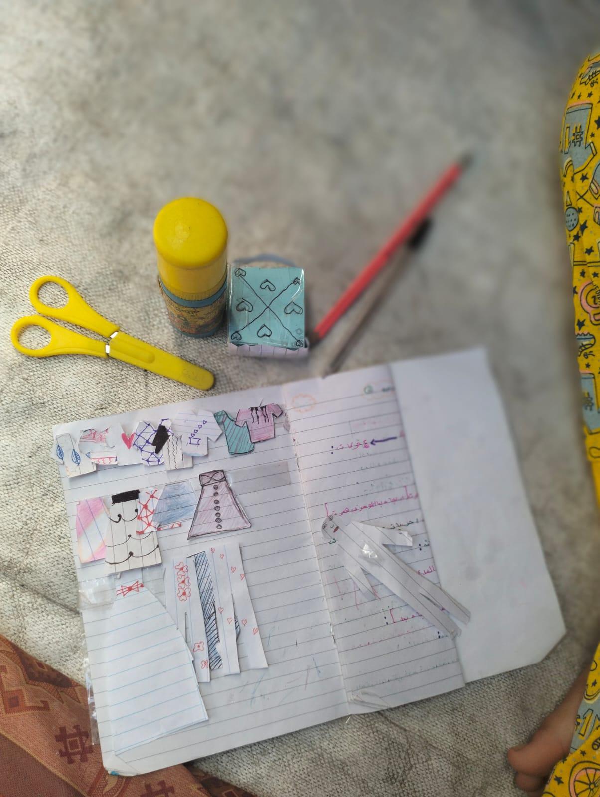 Open notebook page with cut-out paper clothes displayed like a fashion show, accompanied by yellow scissors, glue stick, and a red pencil.