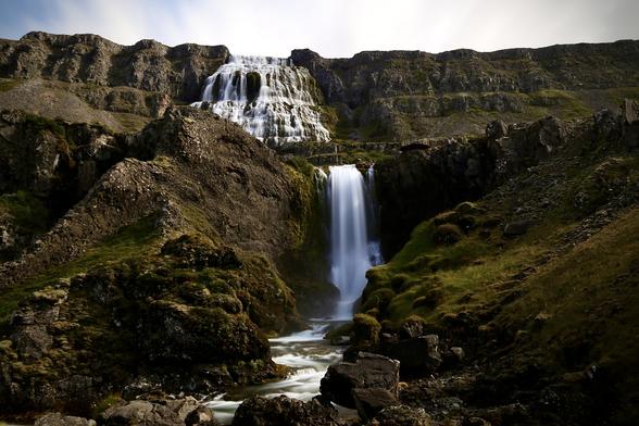 A waterfall cascading down a rugged, moss-covered cliff into a rocky riverbed, surrounded by volcanic terrain.