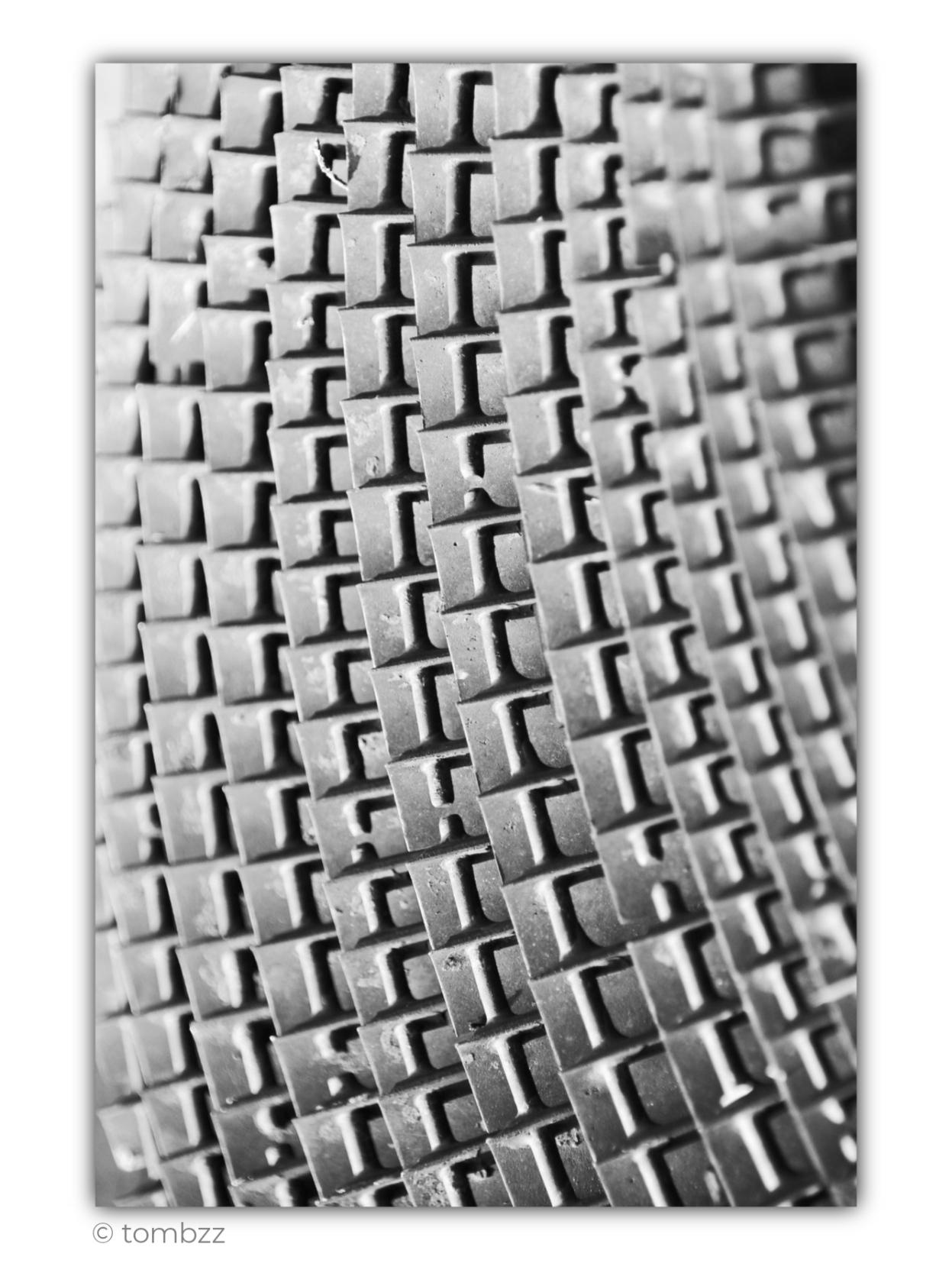Black-and-white photograph taken at the Museum of Coastal Defence on the Hel Peninsula. The frame shows only a small fragment of a large, circular element of an old military machine, most likely dating from the Second World War. Instead of the whole device, the image reveals a dense structure of regularly arranged, massive metal protrusions forming successive, slightly curved rows.
The extreme close-up almost completely strips the object of its original function. Its circular form is reduced to a rhythm of repeating shapes, verticals, recesses, and short horizontal edges. Light falling from the side emphasizes the roughness of the aged metal, its small imperfections and surface irregularities, while the deep shadows between the elements enhance the sense of depth.
The central part of the frame remains the sharpest. Toward both edges, the structure gradually loses clarity, so the repeating elements begin to dissolve into the background. The shallow depth of field, tight cropping, and pe