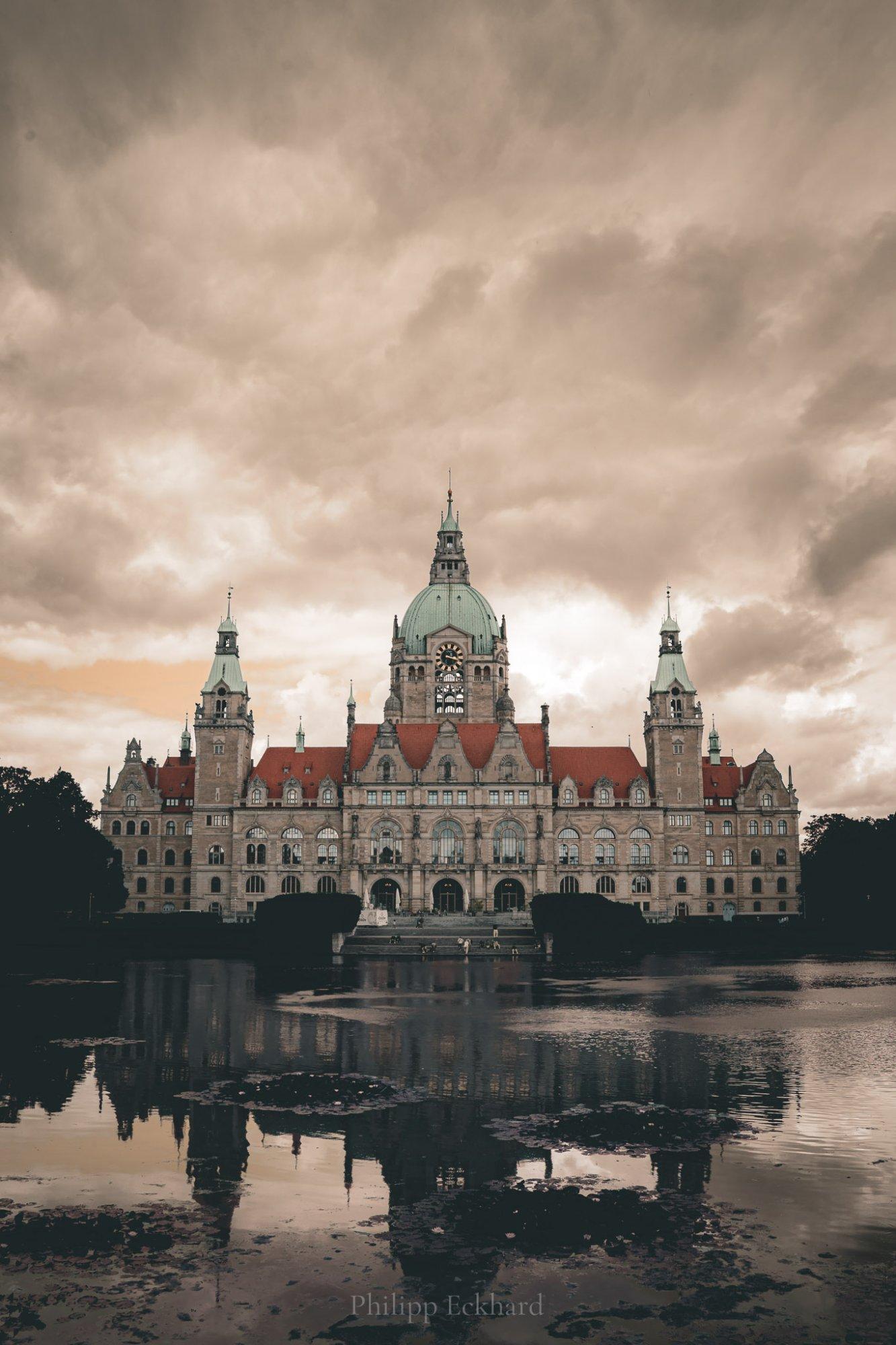 Neues Rathaus Hannover mit seiner markanten Kuppel unter dramatischem Wolkenhimmel, gespiegelt im Wasser des Maschparks.

Hanover New Town Hall with its distinctive dome beneath a dramatic cloudy sky, reflected in the water of Maschpark.