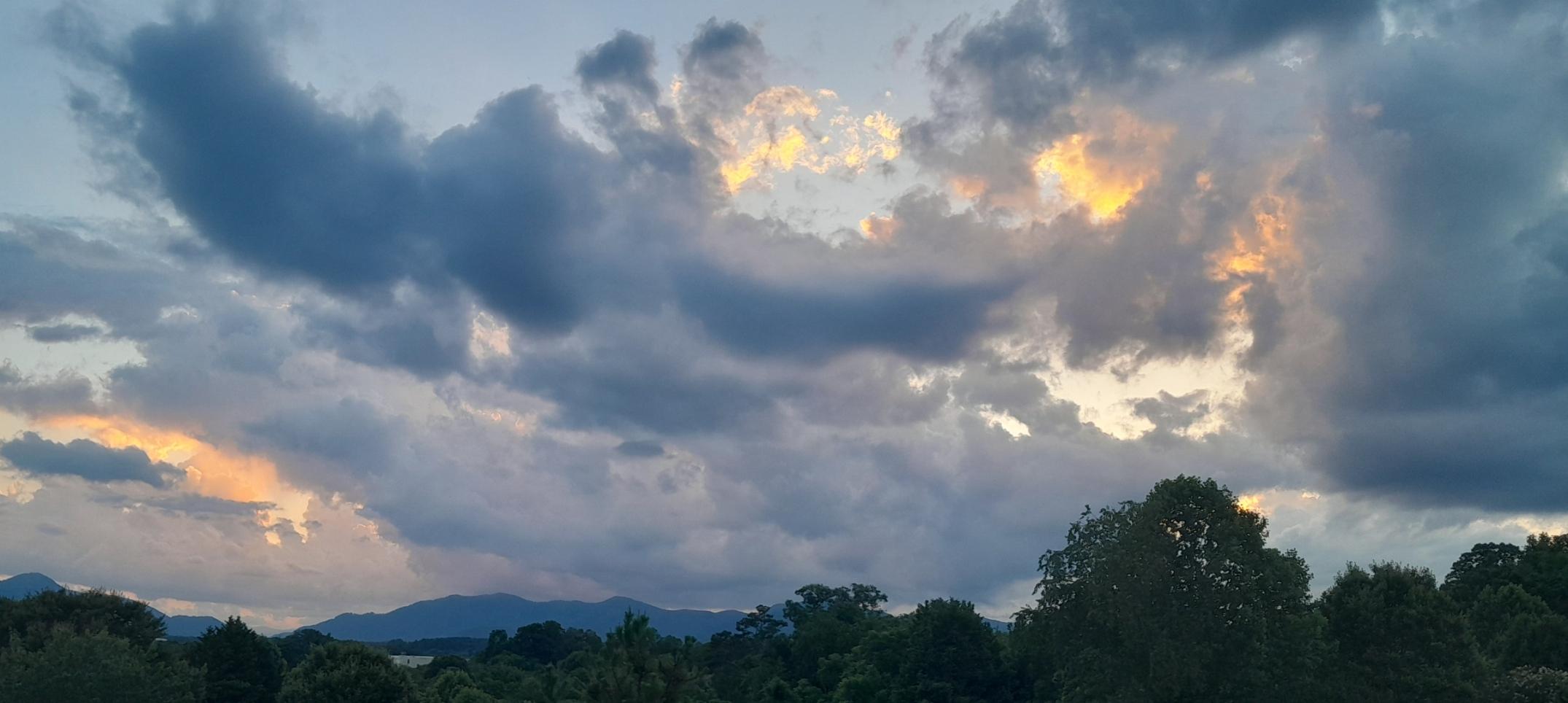 Dark vegitation and trees foreground, mountains in the distance with cloud covered skies hilited in orange by the setting sun