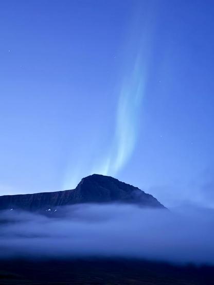 A plume of green aurora rises above a mountain shrouded with clouds